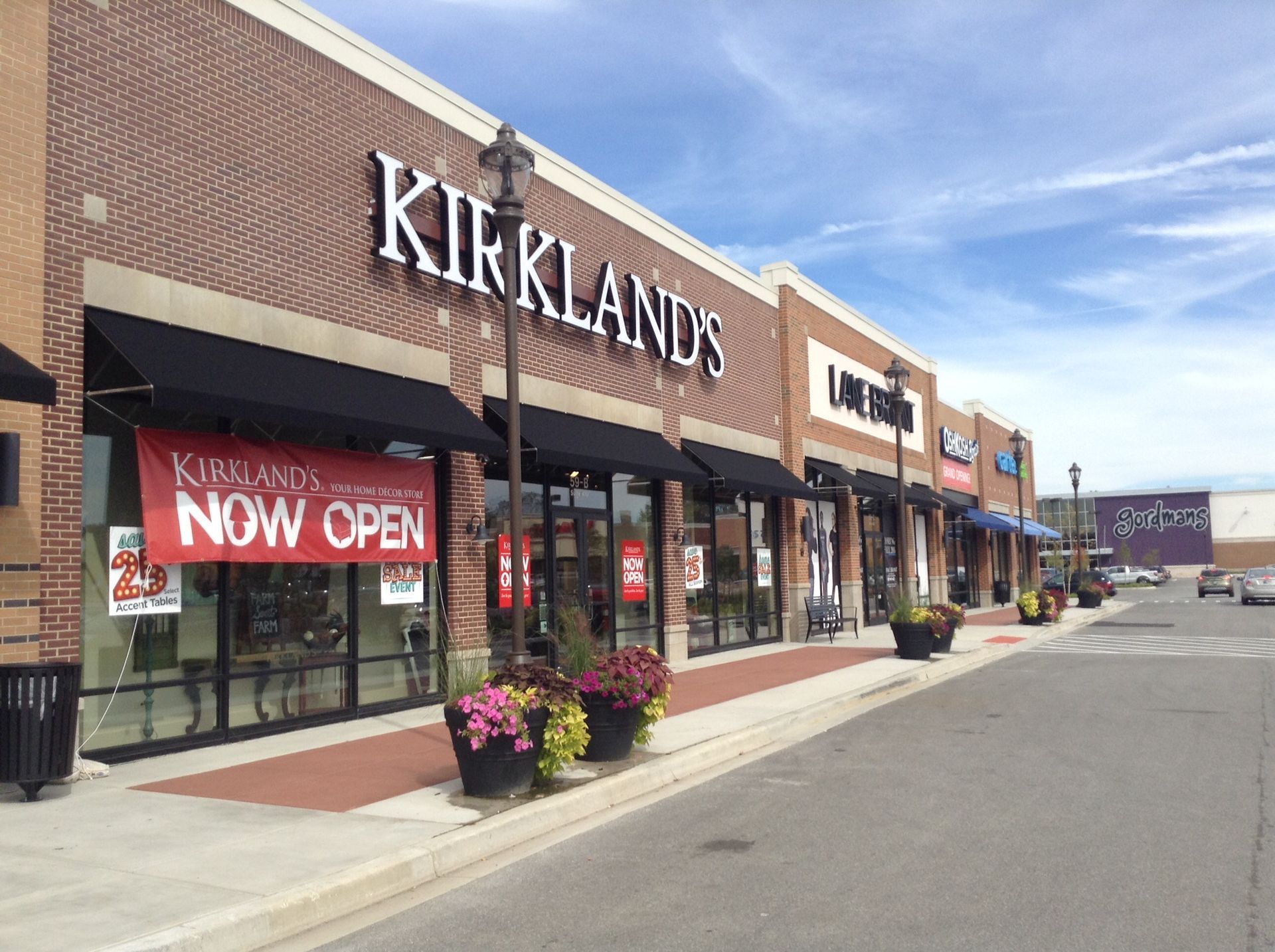 Kirkland’s storefront in a strip mall with a red “NOW OPEN” sign and flower planters on the sidewalk