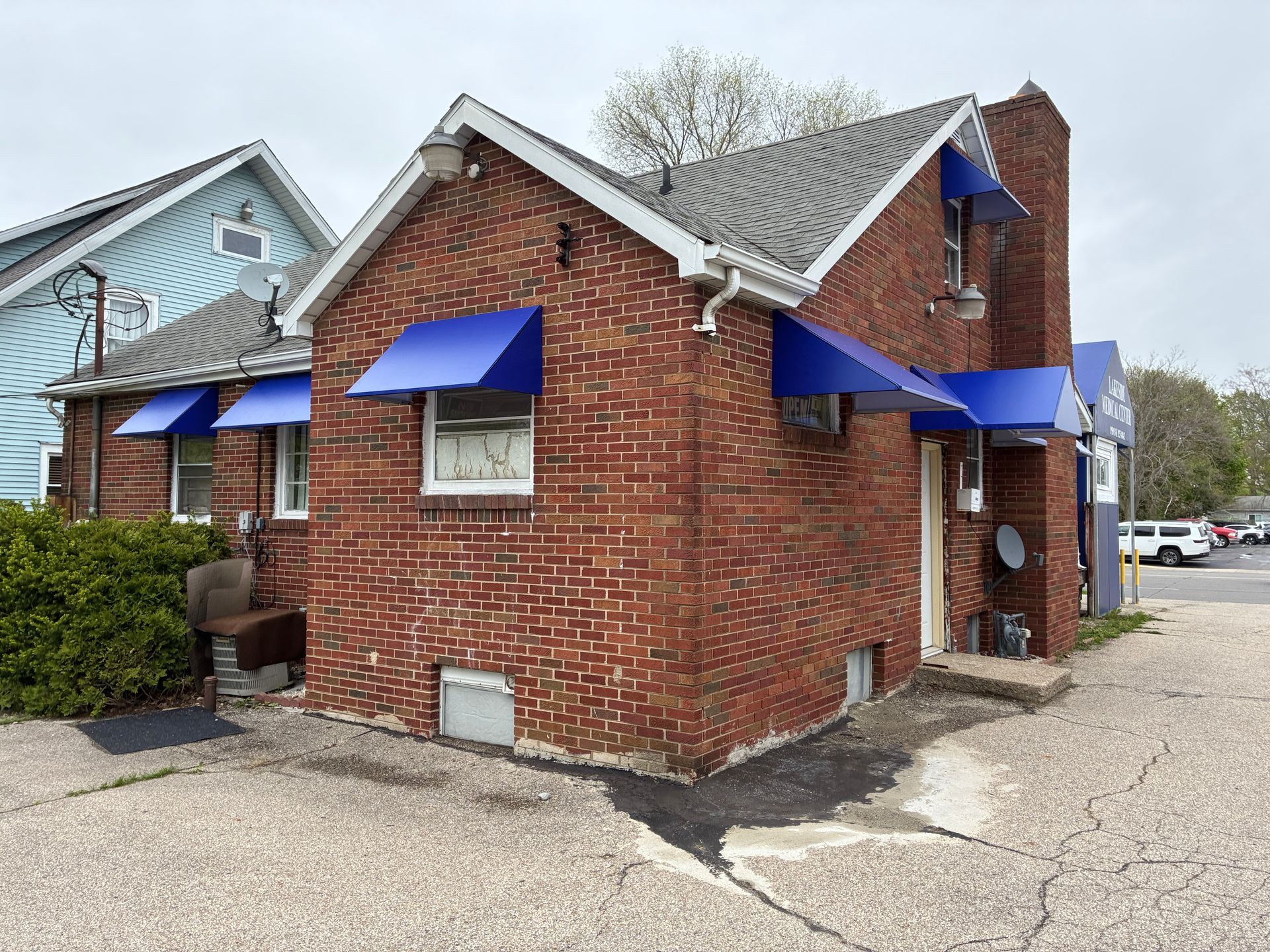 Small red-brick house with blue awnings and white trim on a gravel lot