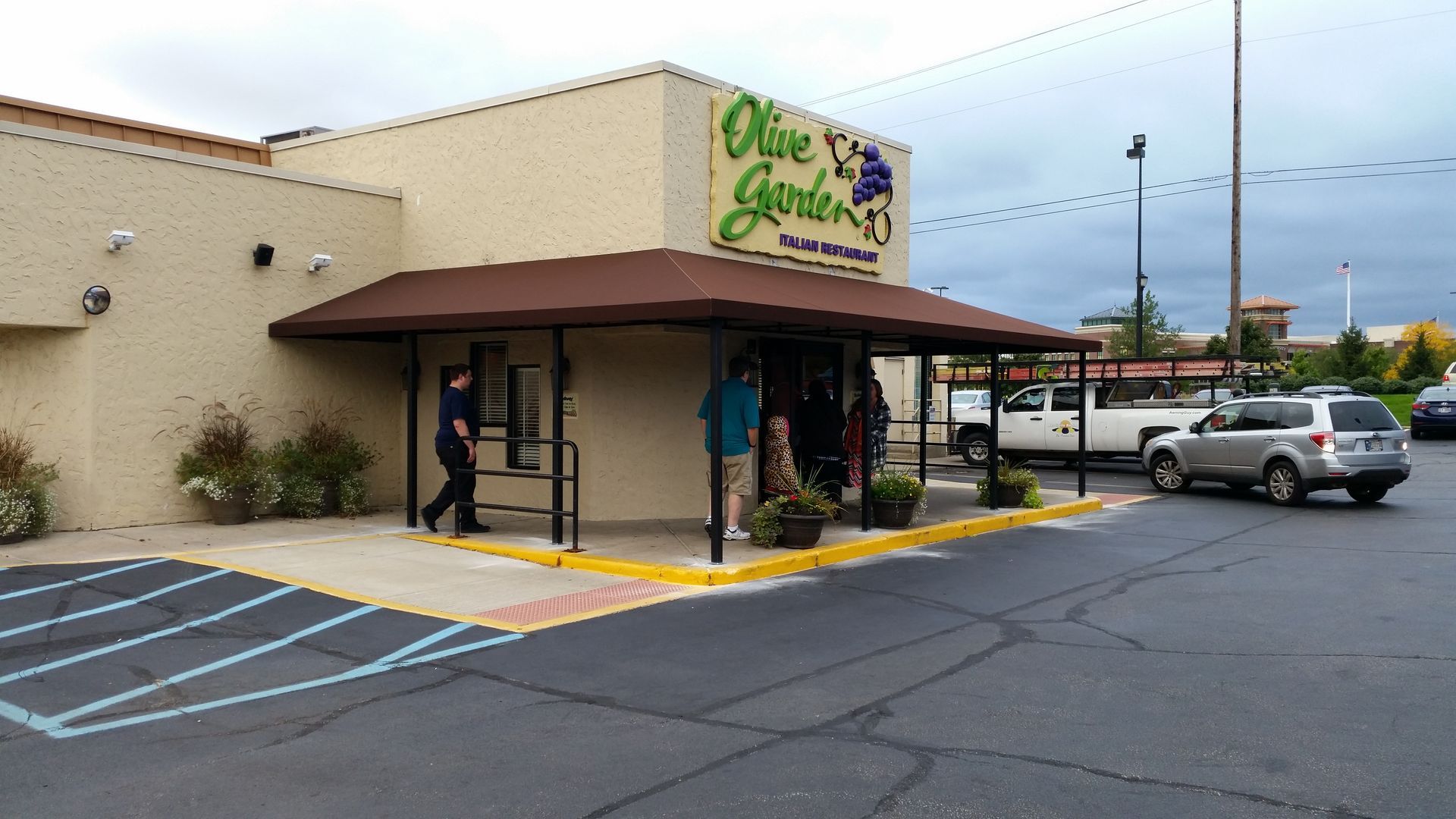 Storefront with brown awning and yellow curb, a few people walking by, and a parking lot with cars.