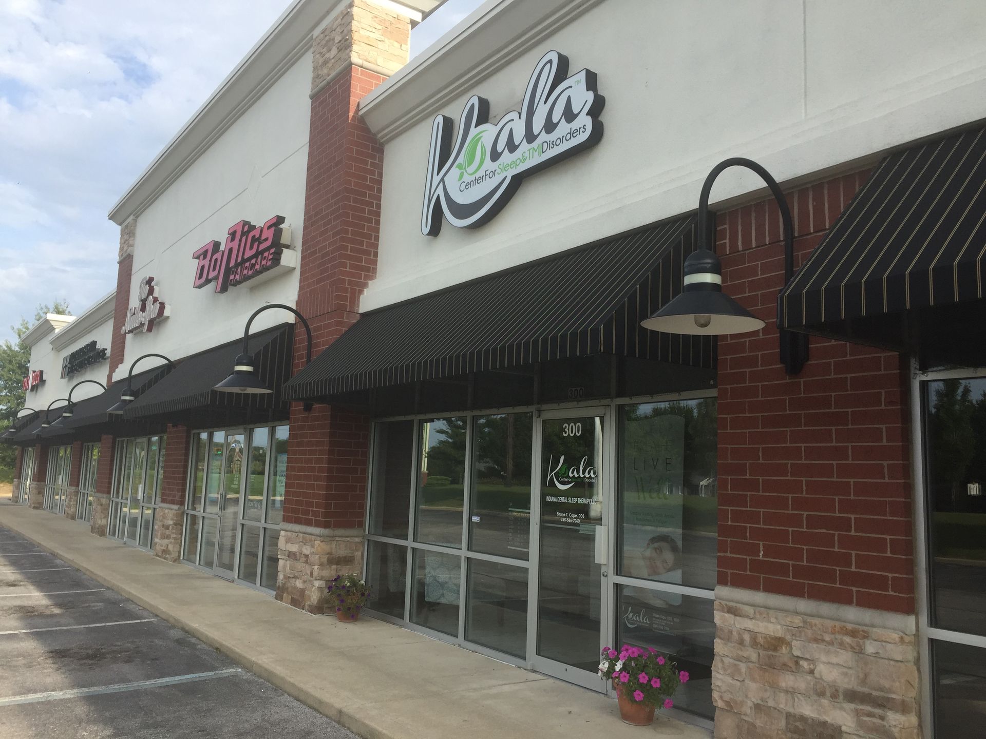Strip mall storefronts with striped awnings, glass doors, and brick accents on a sunny sidewalk.