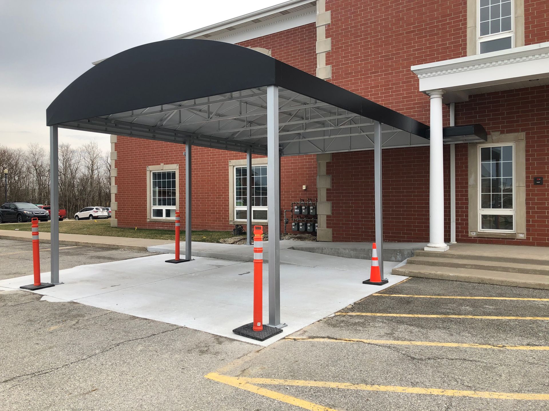 Covered parking canopy beside a red brick building, with orange bollards and accessible parking spaces.