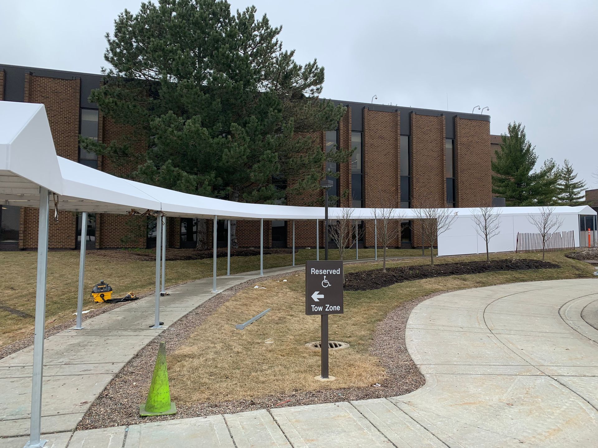 Covered walkway beside brick building with white tents, a tree, and a paved path under construction.