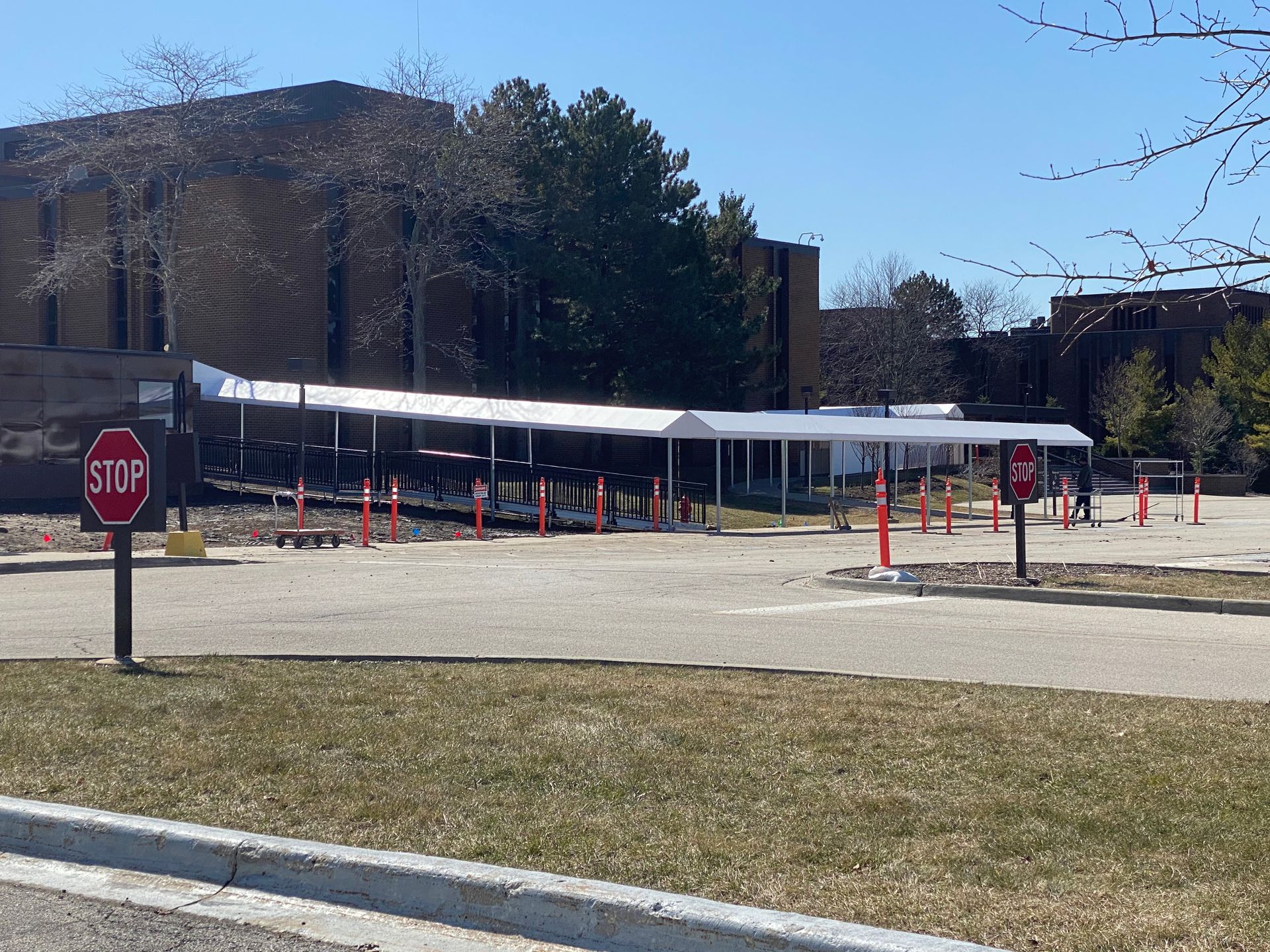 Campus sidewalk covered by a long white canopy, lined with orange cones and a stop sign in front.