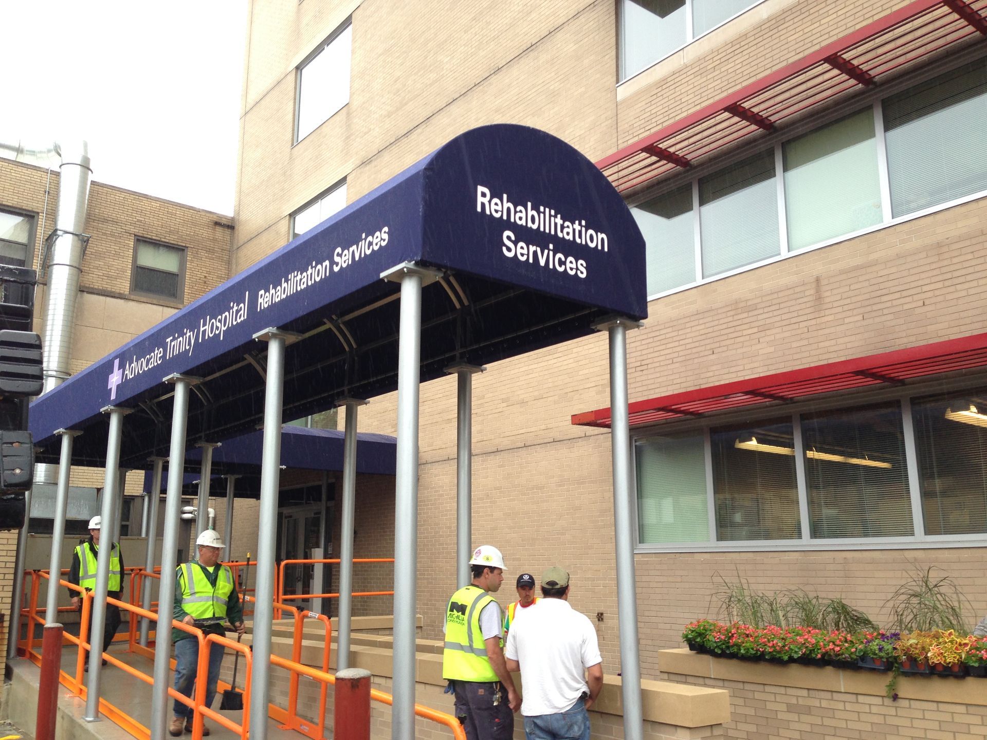 Workers under scaffolding outside a building with a blue awning labeled “Rehabilitation Services”