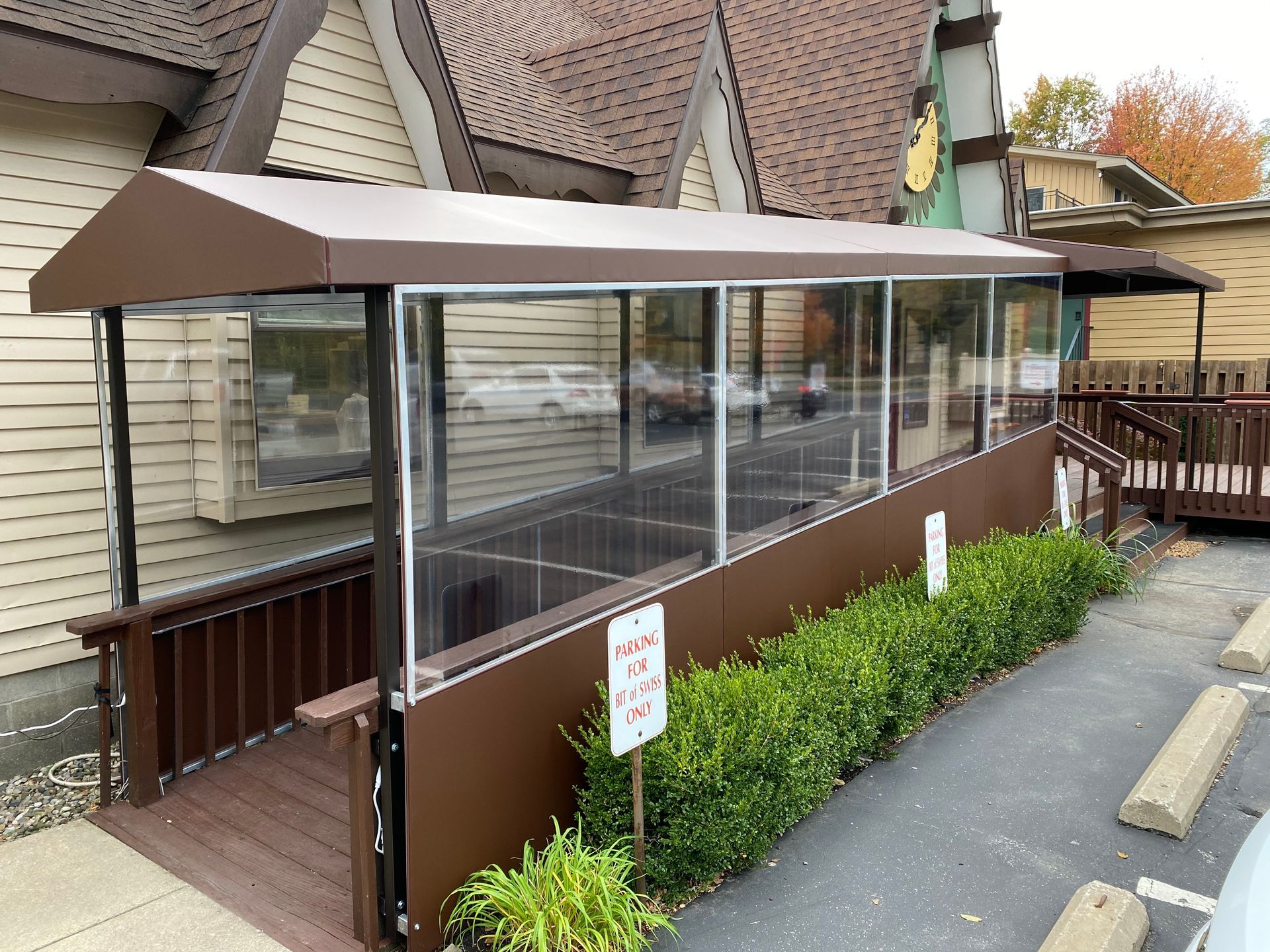 Covered outdoor walkway with clear panels and brown trim beside a building and hedge-lined path