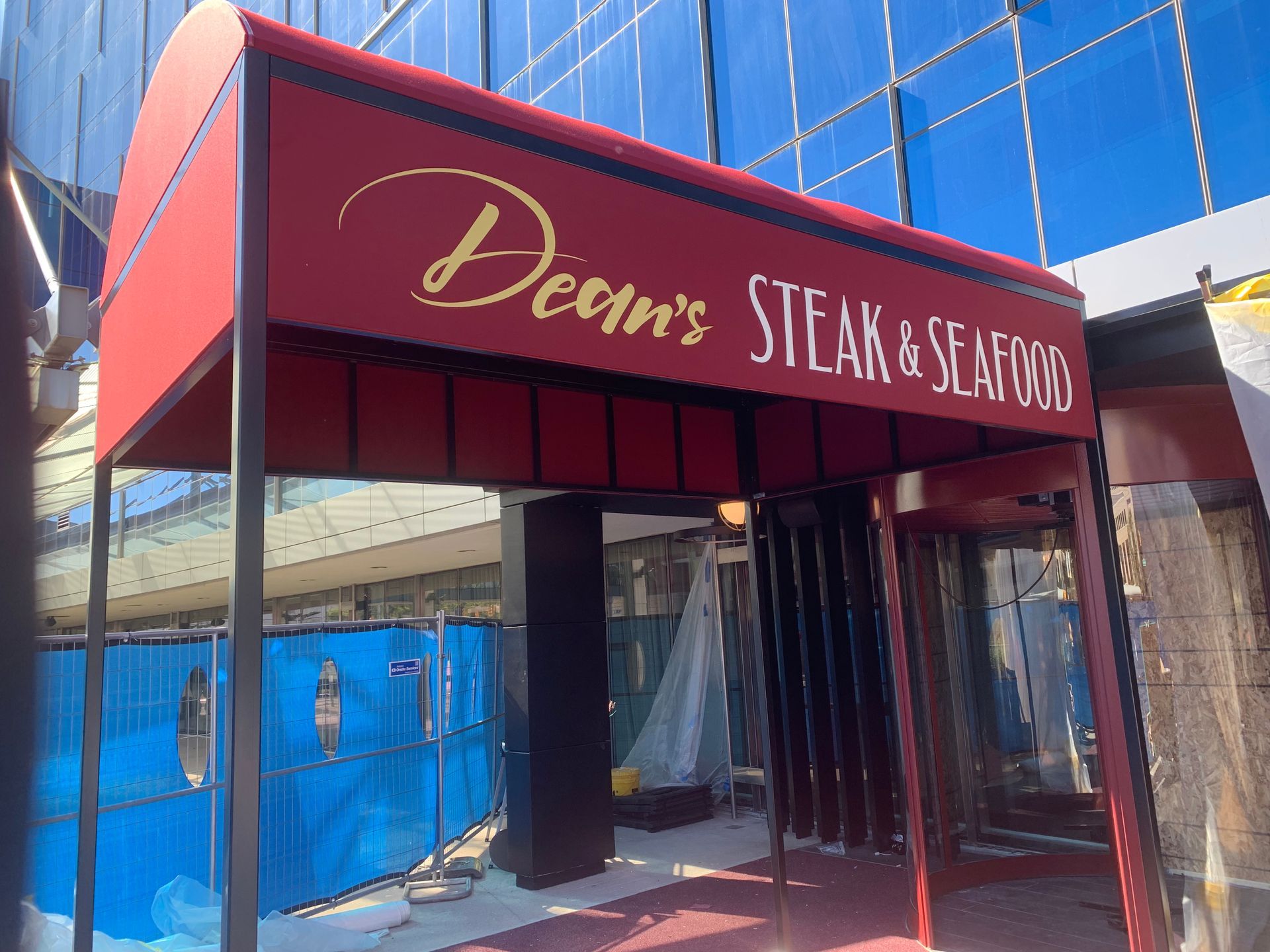 Restaurant entrance with a red awning reading “Dean’s Steak & Seafood” and blue construction fencing nearby