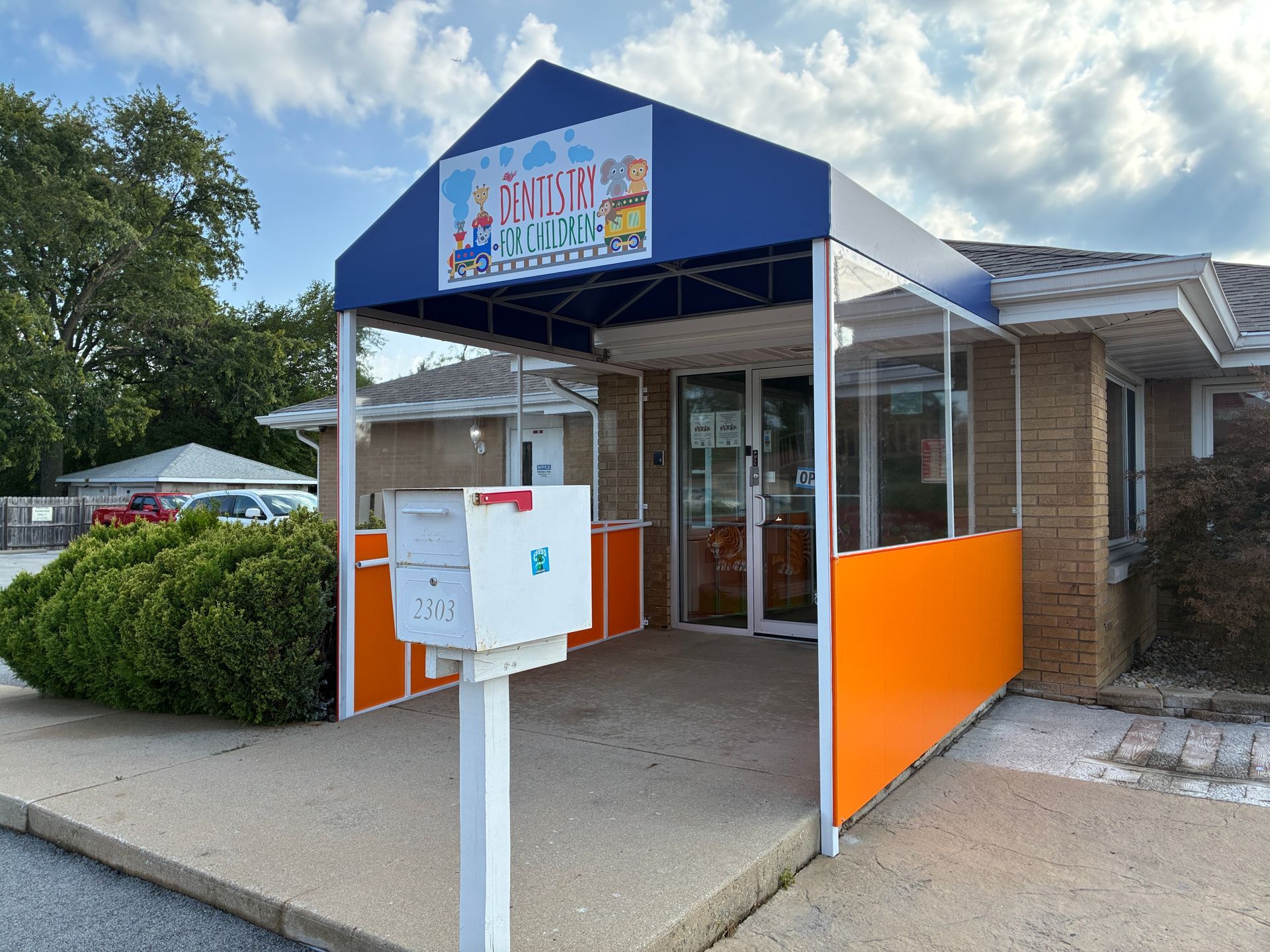Entrance to a building with blue awning, orange panels, and a white drop-off box on the sidewalk.
