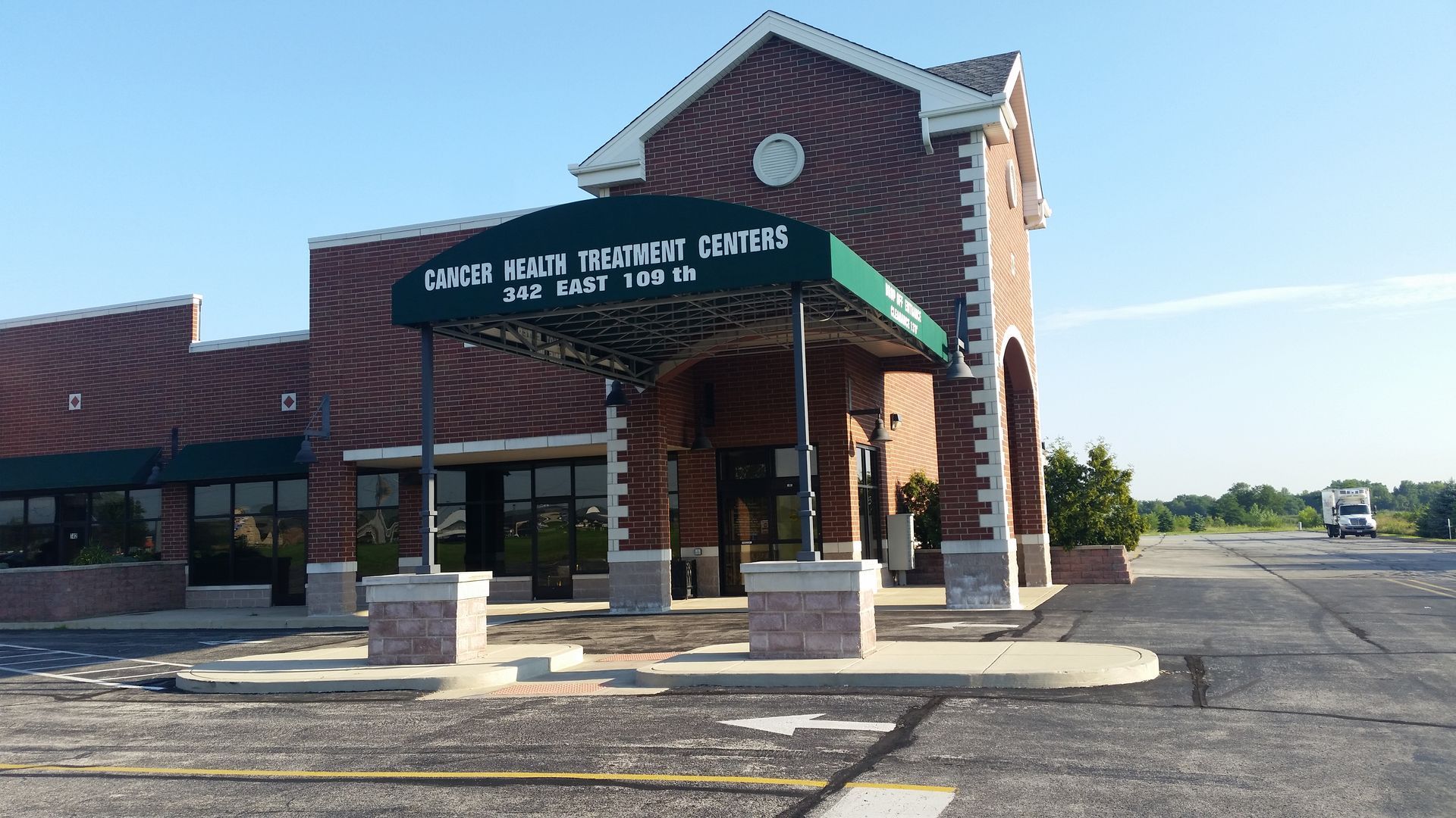 Brick school entrance with green awning and covered drop-off area, viewed from parking lot.