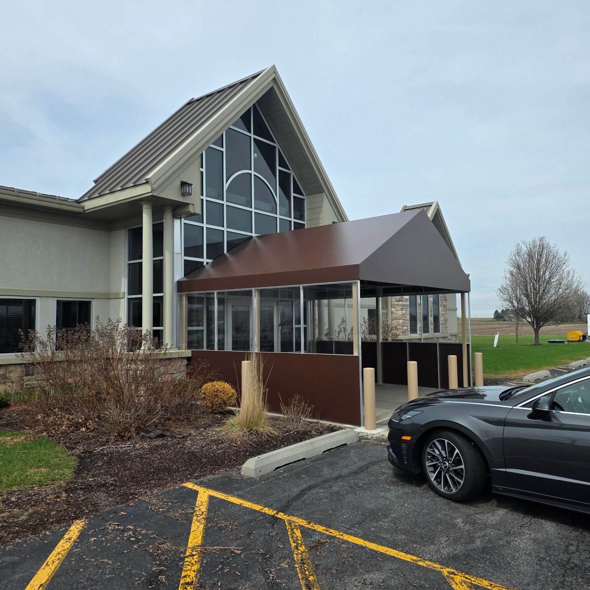 Church entrance with brown awning, glass windows, and a parked car in a lot on an overcast day
