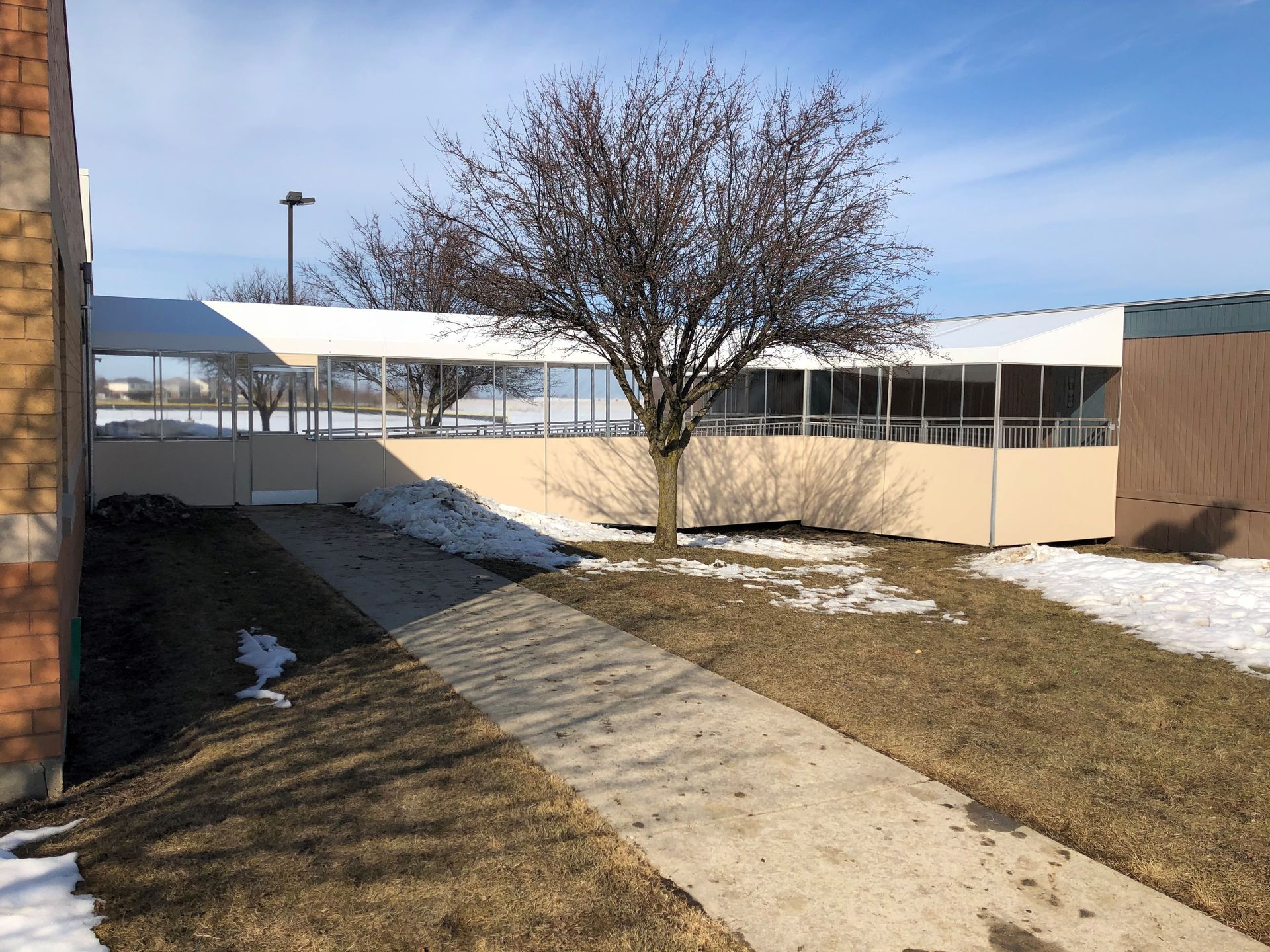 Snowy courtyard with a concrete path, bare tree, and beige building under a clear blue sky