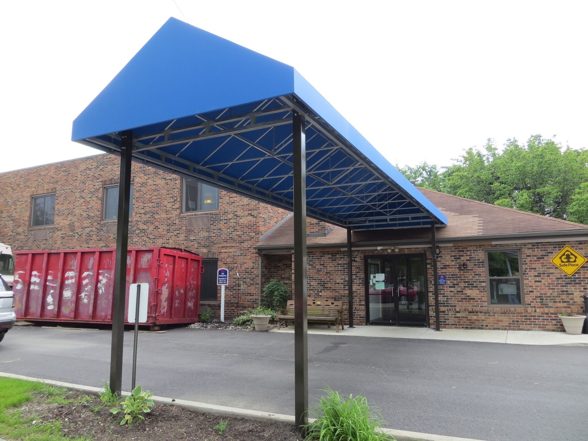 Blue canopy over brick building entrance with red dumpsters and parked cars outside