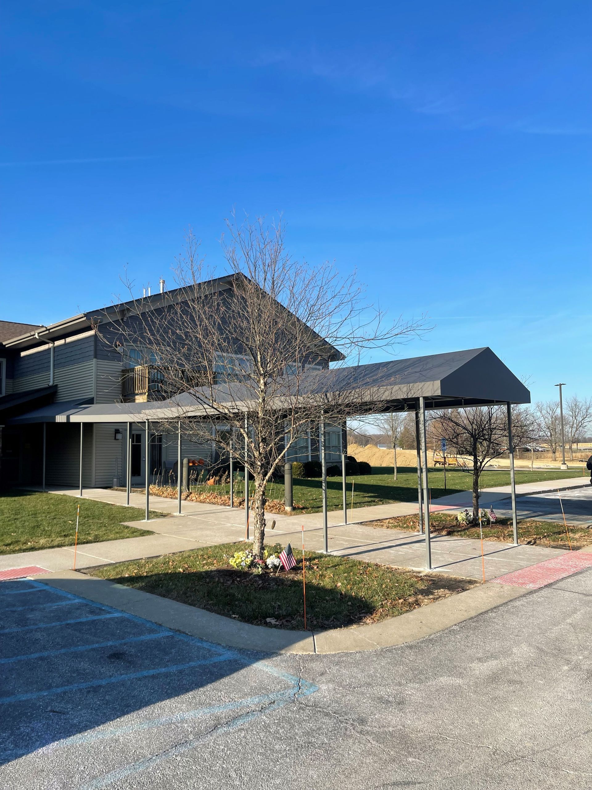 Covered walkway beside a modern building, with a young tree, blue sky, and parking lot foreground