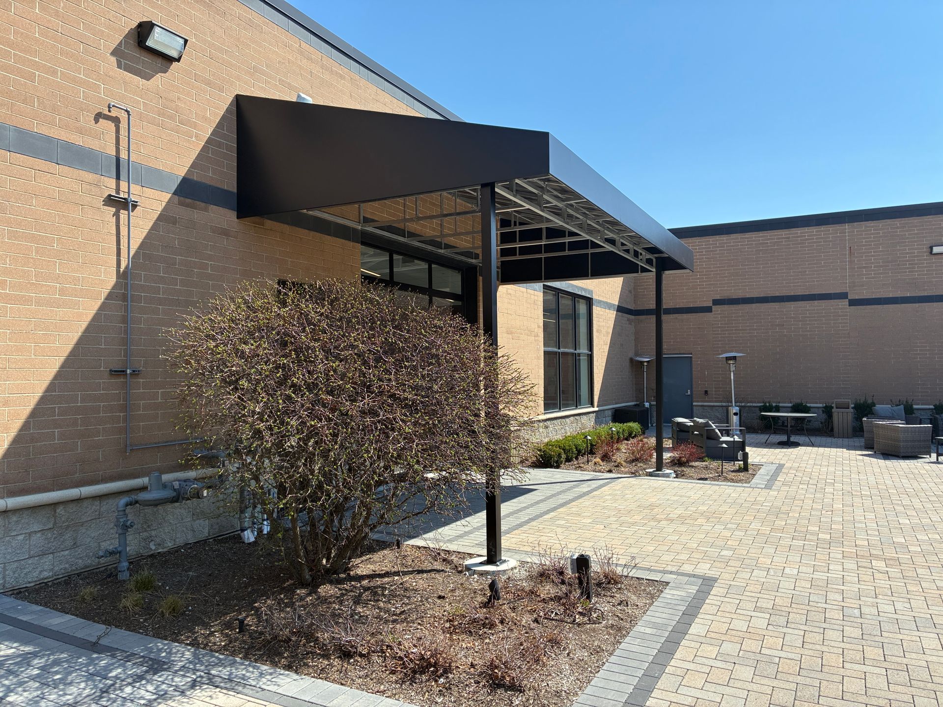 Building entrance with black awning, brick walls, shrubs, and a paved walkway under clear blue sky