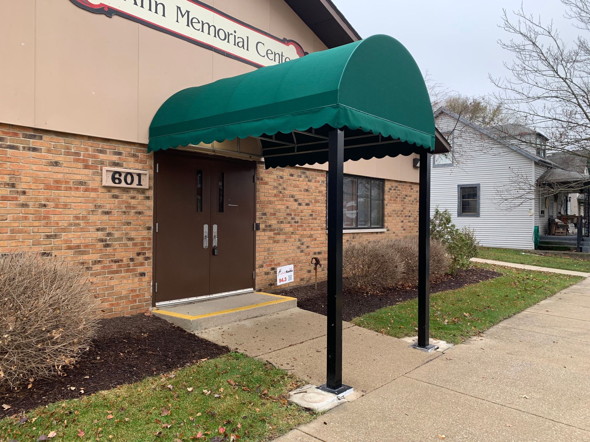 Building entrance with green awning, brick wall, and concrete walkway outside a memorial center