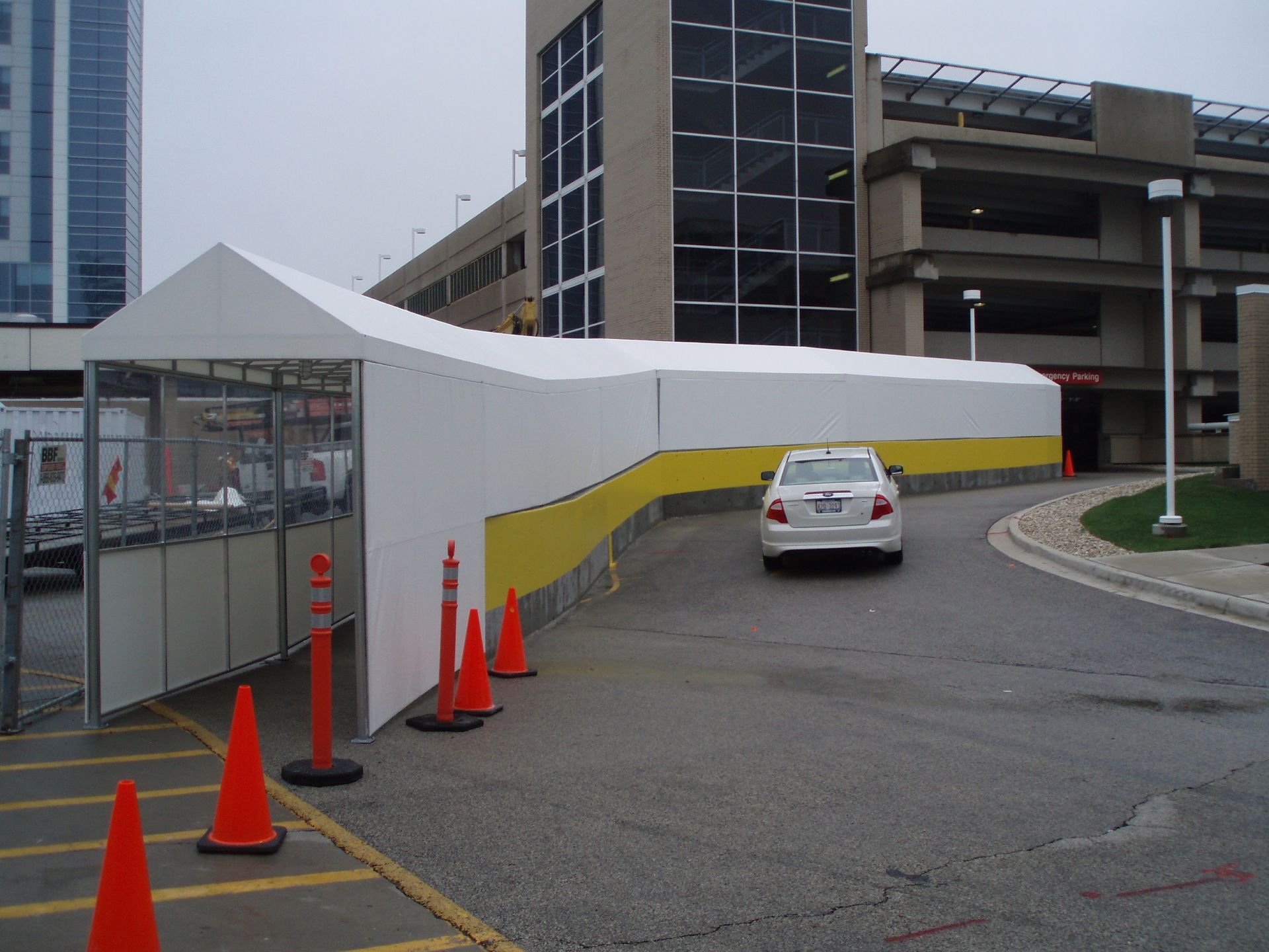 White tent barrier beside a parking lot, with orange cones and a white car on a curved road.