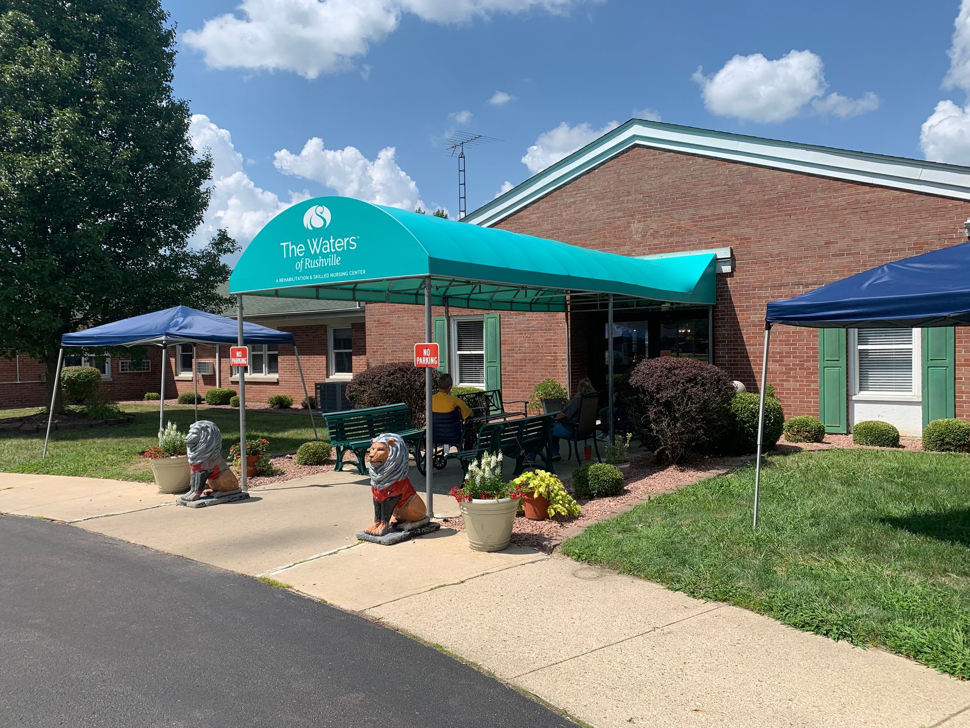 Outdoor entrance with turquoise canopy, potted plants, and chairs beside a brick building on a sunny day