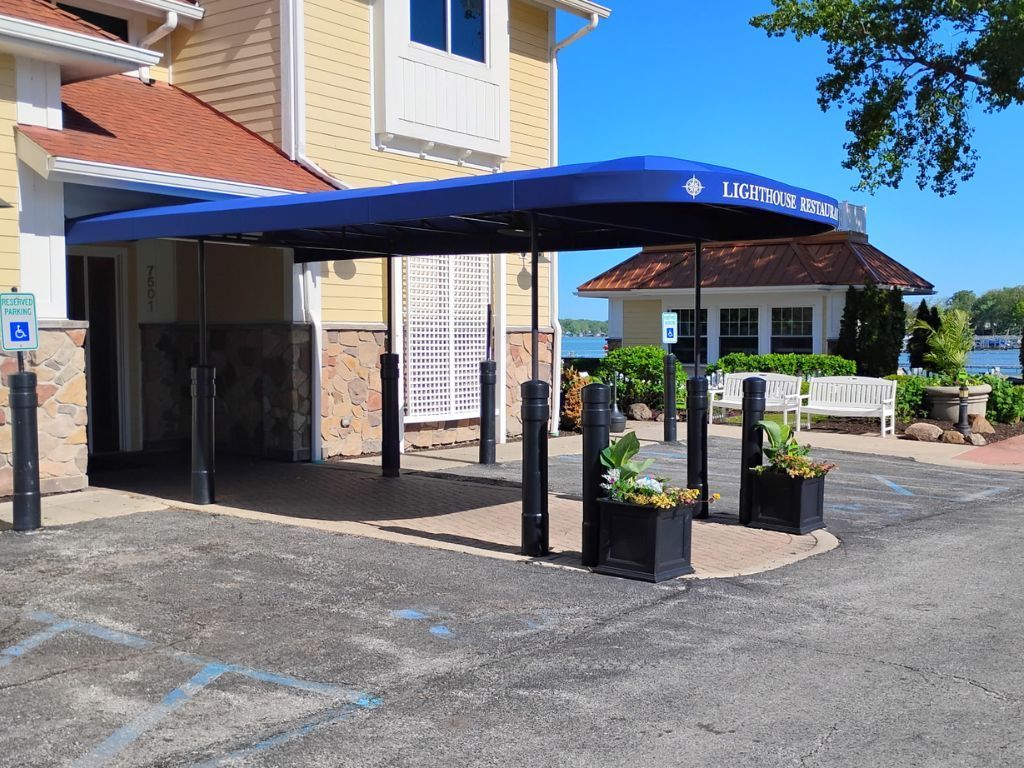Covered hotel entrance with blue awning, parking area, and potted plants by the building.