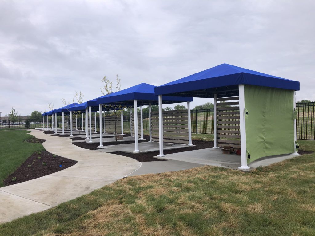 Covered walkway with blue roofs and white posts along a paved path in a grassy park.