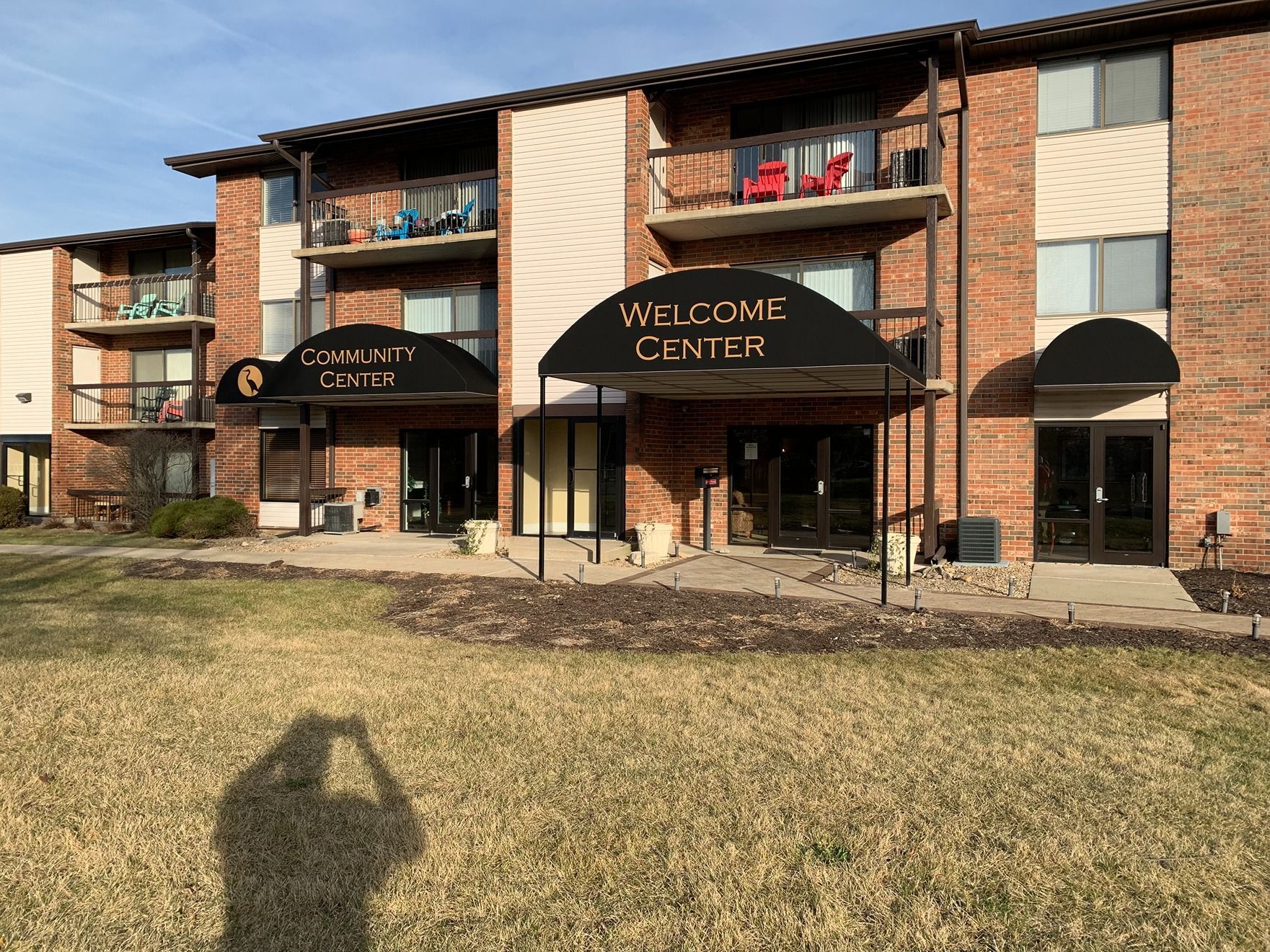Apartment-style building with a welcome center entrance and covered walkway, set on a grassy lawn.