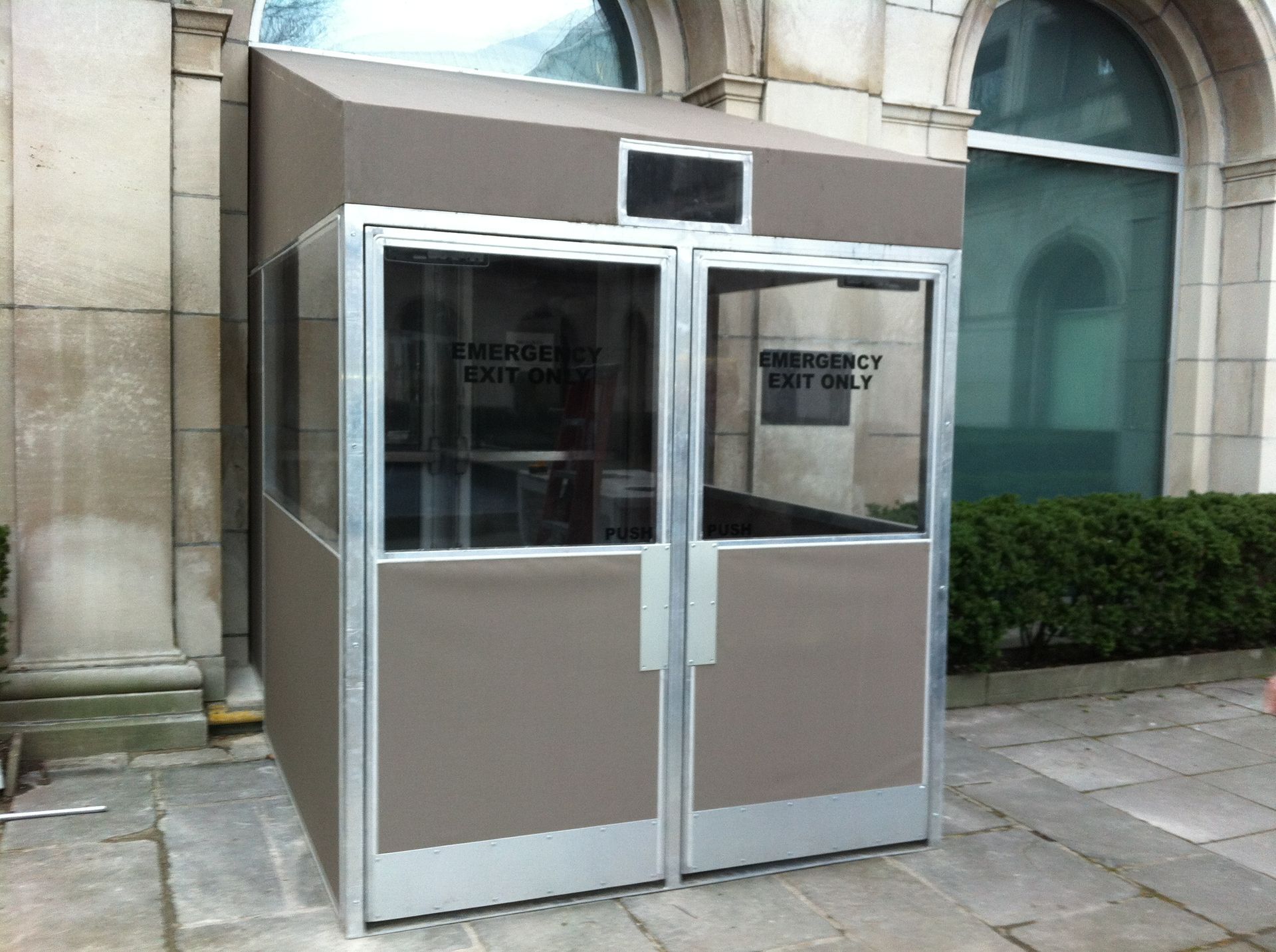 Small accessible kiosk with glass double doors and silver panels on a stone walkway beside a building