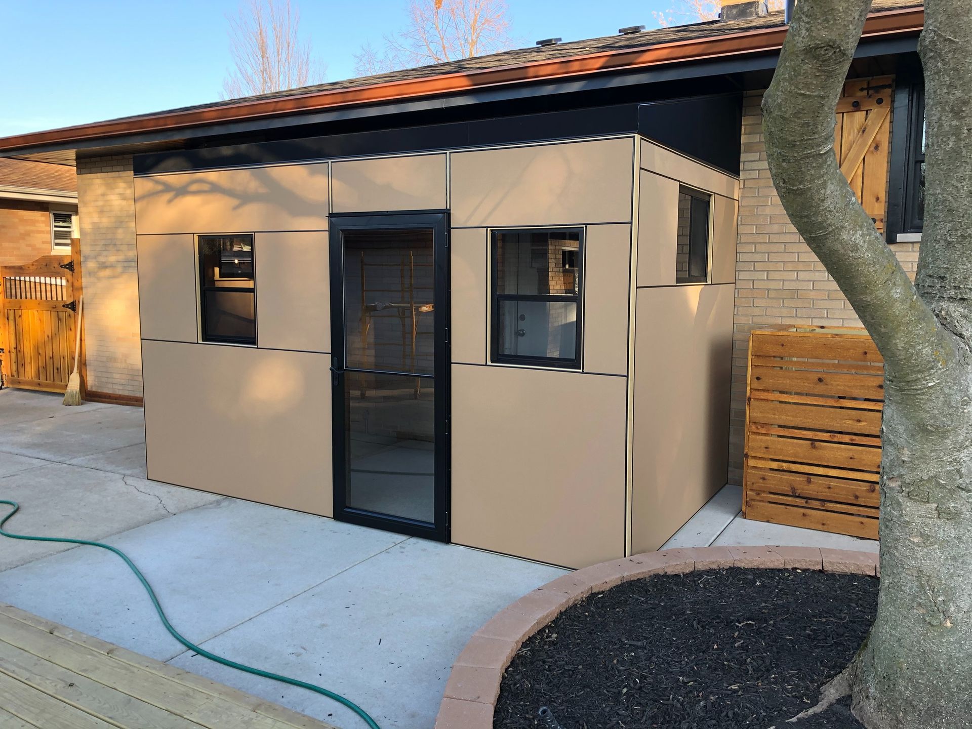 Small beige outdoor shed with black-framed windows and door beside a tree and wooden fence.