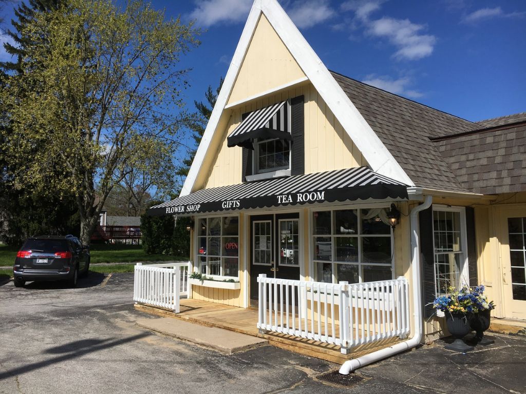 Small yellow house with white trim, porch railing, black awnings, and a parked car in the driveway.