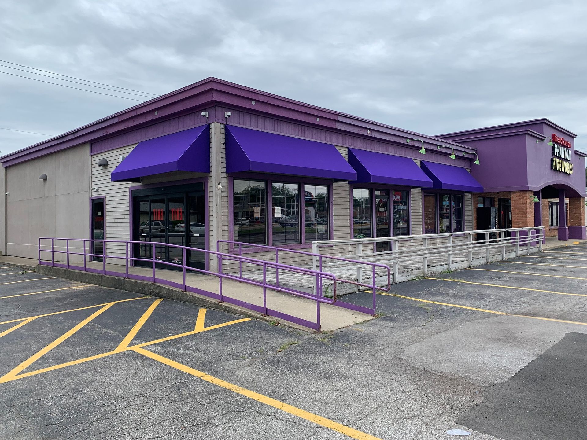 Corner storefront with purple awnings, metal railing ramp, and empty parking lot.