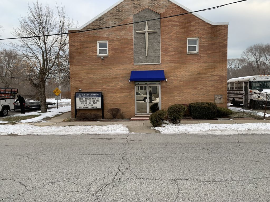 Brick church building with a blue awning and cross, seen across a snowy street.