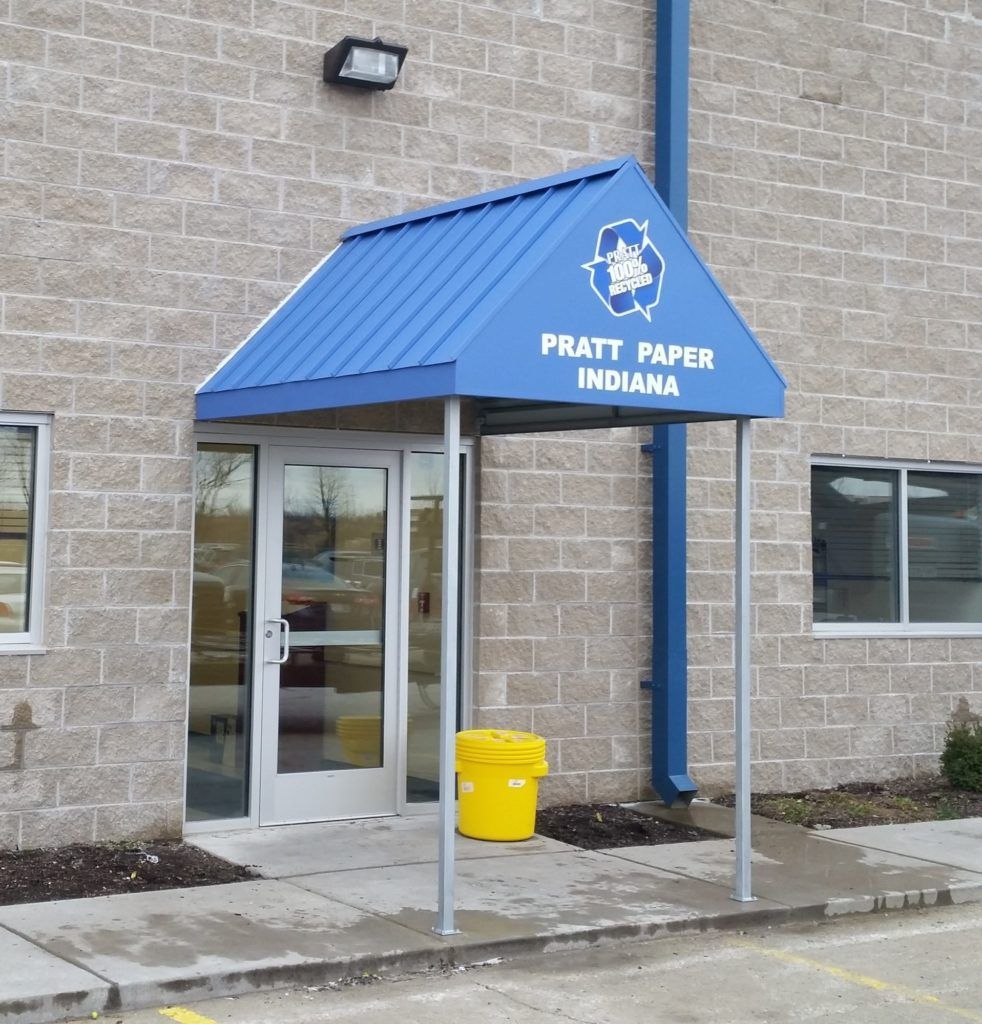Blue awning entrance labeled “PRATT PAPER INDIANA” on a brick building, with a yellow trash can nearby.