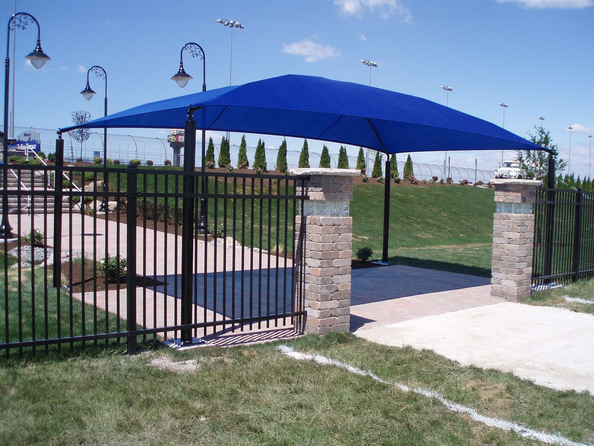 Blue shade canopy over a gated walkway beside a grassy park and stone pillars under a clear sky.