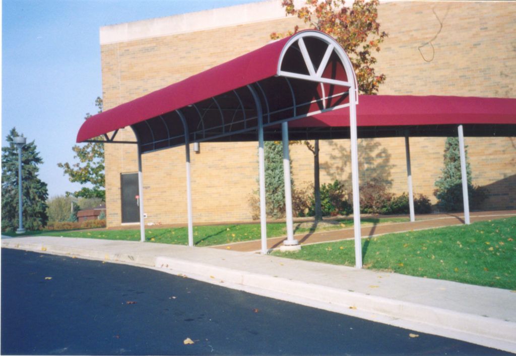 Building entrance with a maroon awning, white posts, and a sidewalk beside a grassy lawn