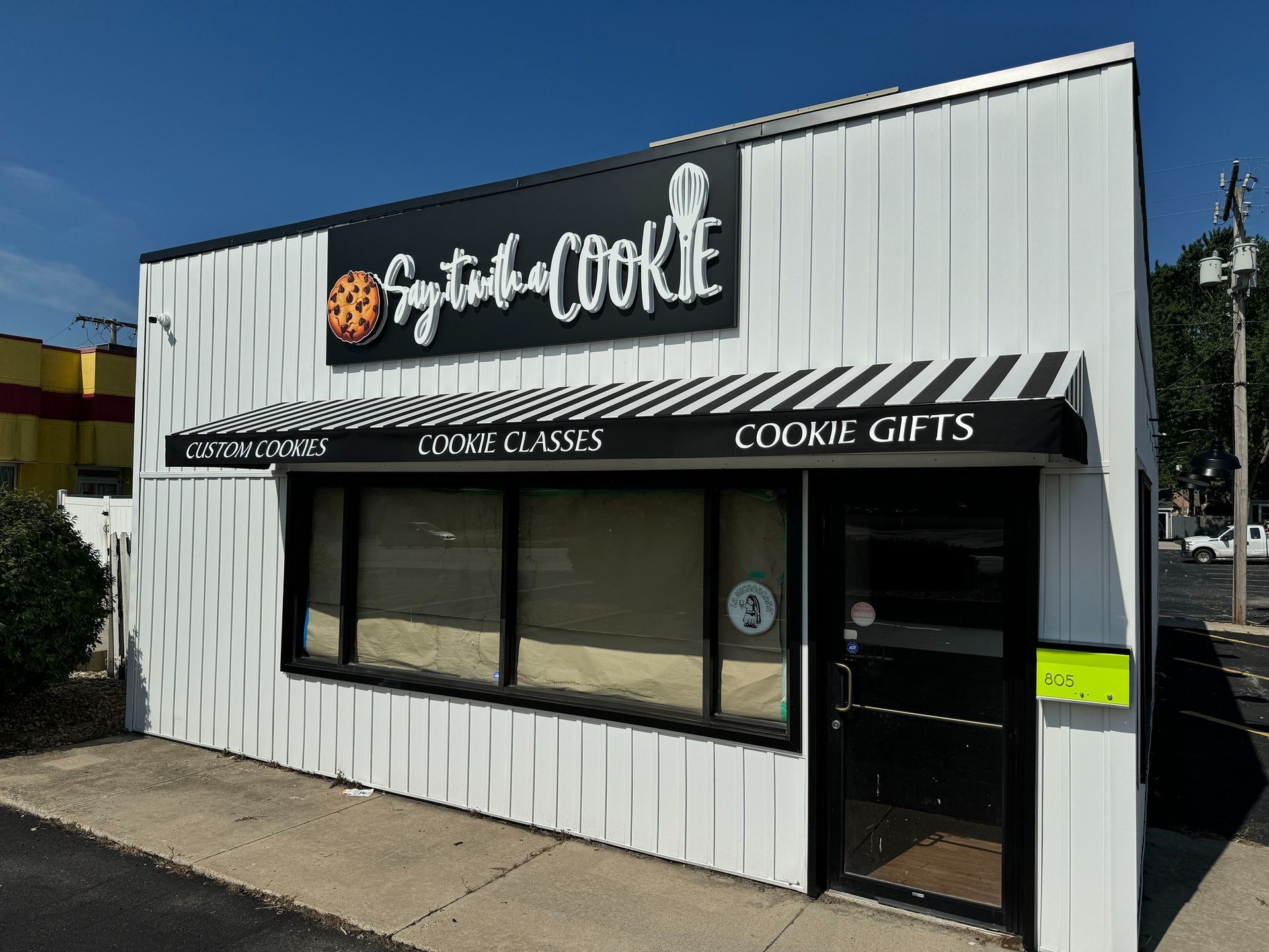 Small white storefront for a cookie shop with black sign and awning, large front windows, and open door
