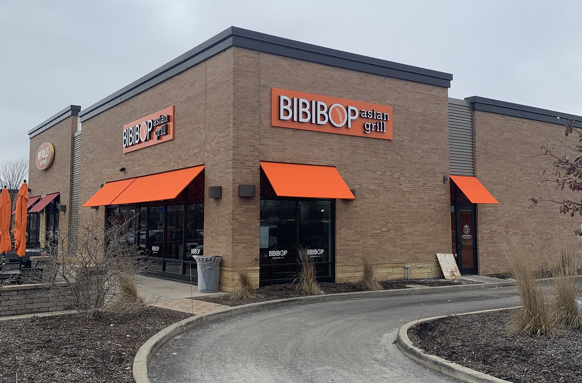 Corner view of a BIBIBOP storefront with orange awnings and a curved sidewalk in front