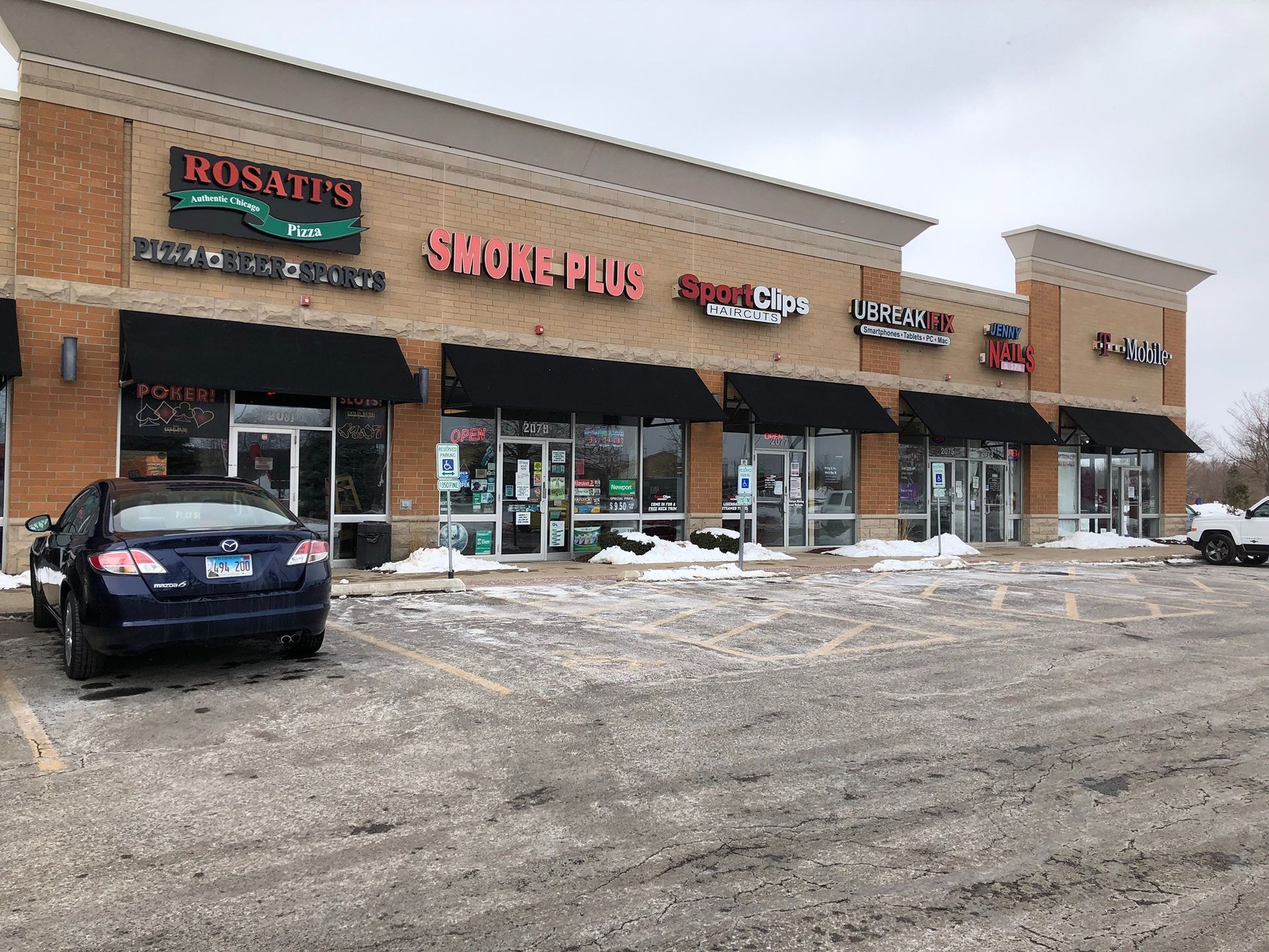 Snowy strip mall storefronts with parked cars and a beige facade
