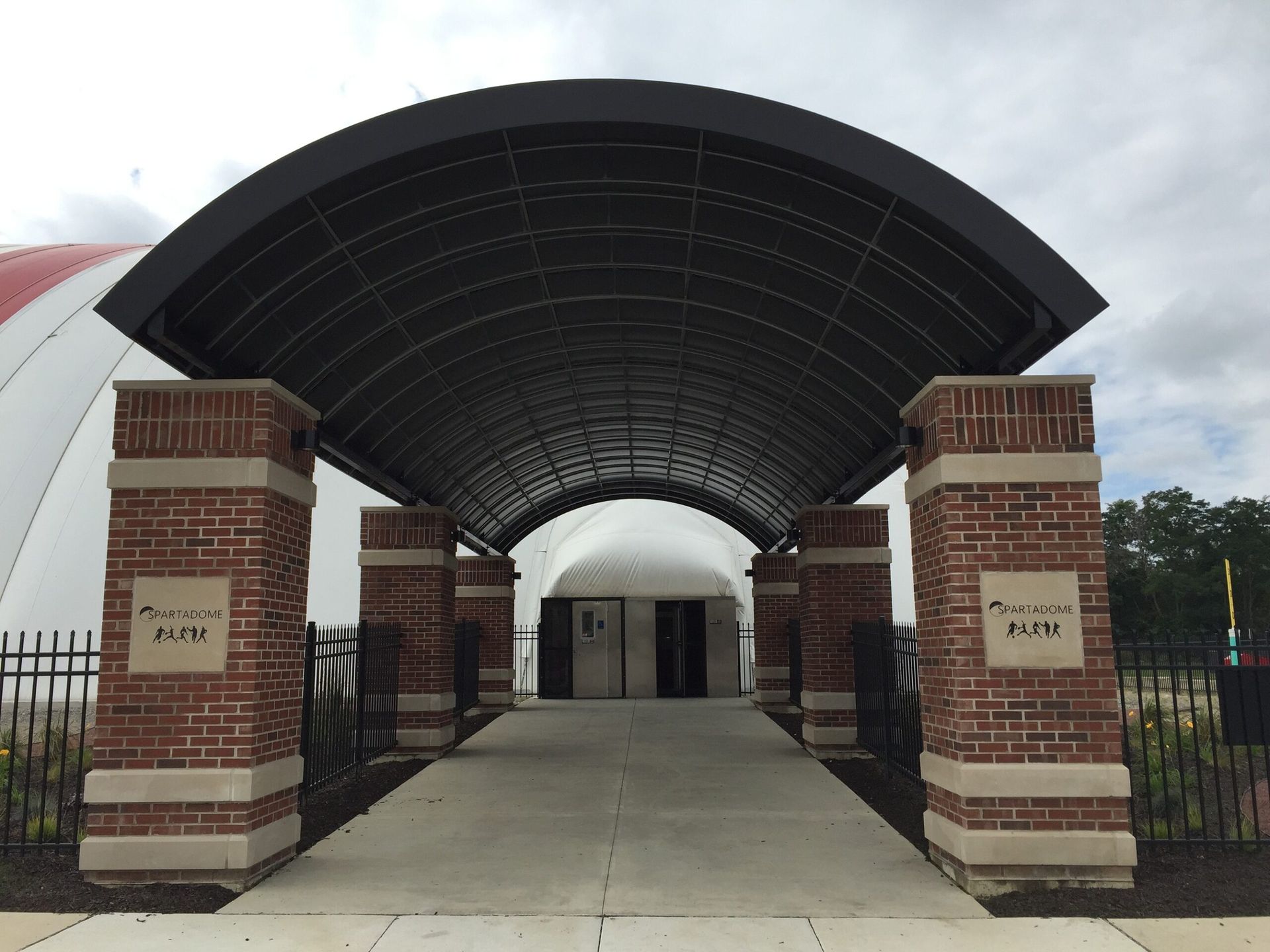Covered brick entrance gate with arched black canopy leading to a building, viewed from a walkway