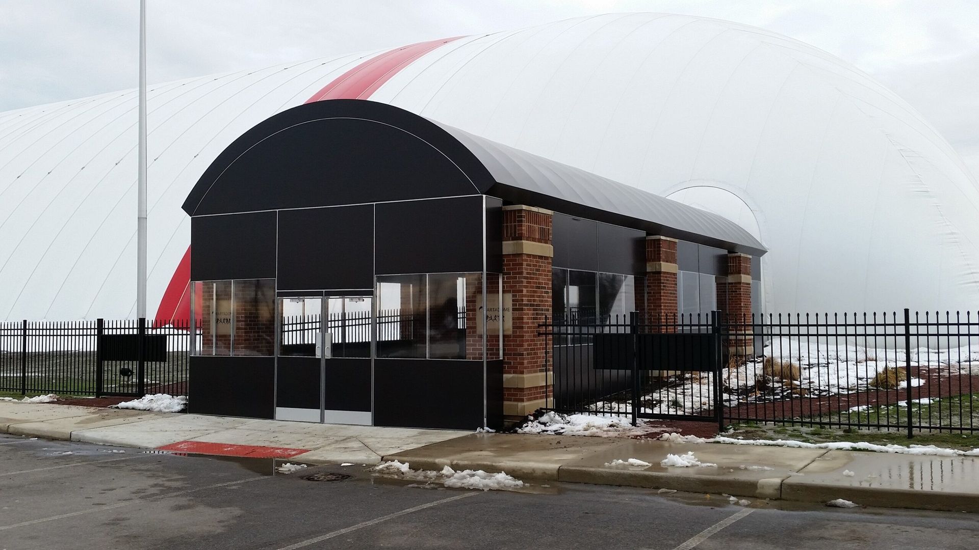 Small modern building with a curved black canopy and fenced entrance, covered in snow.