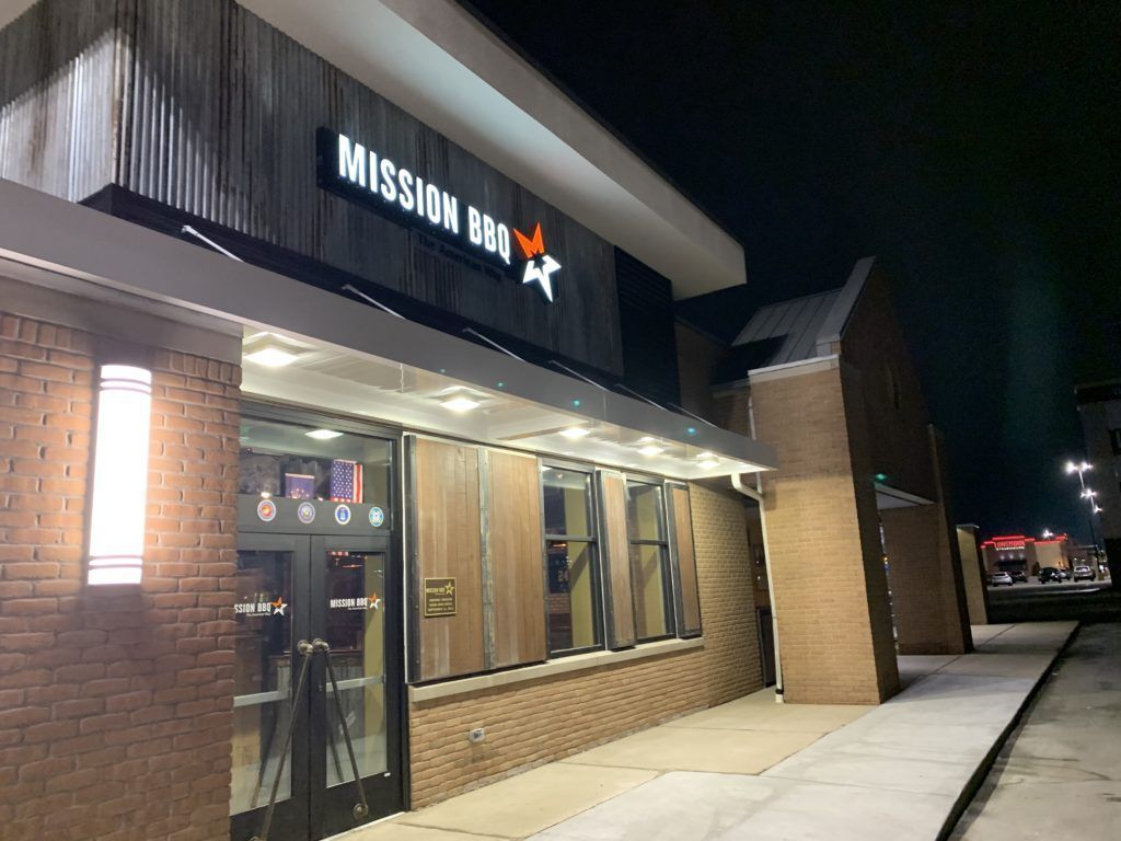 Mission BBQ storefront at night, with lit windows, brick exterior, and sidewalk in front.