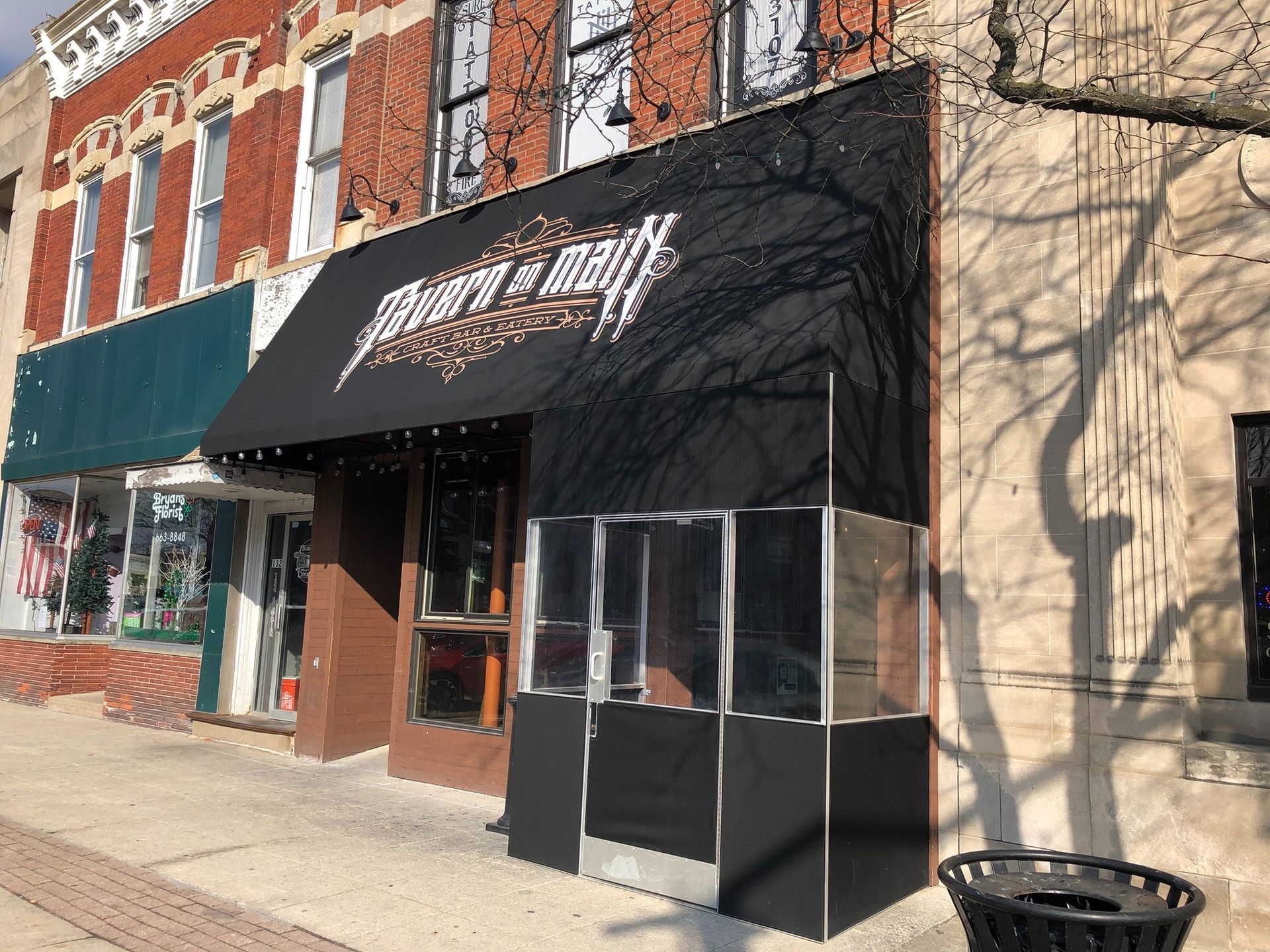 Storefront with black awning on a brick building along a sunny street, with a trash bin on the sidewalk