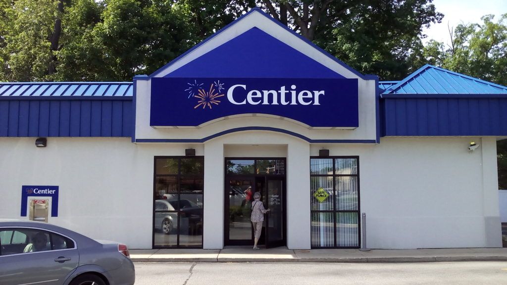 Centier bank storefront with blue sign and entrance, parked car at left, trees behind