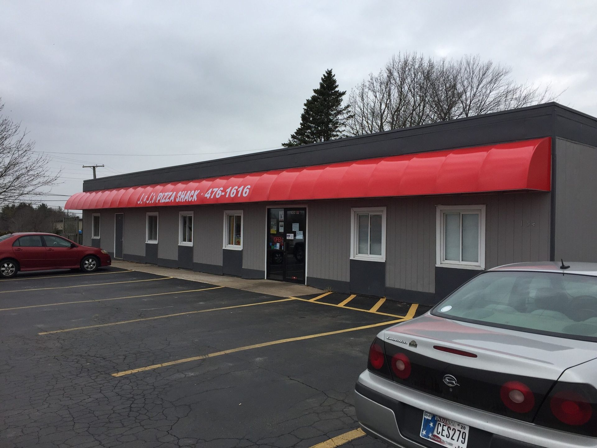 Small gray storefront with a red awning and parked cars in front on an overcast day