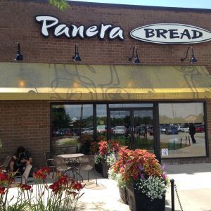 Panera Bread storefront with outdoor seating, brick facade, and flower planters on a sunny day