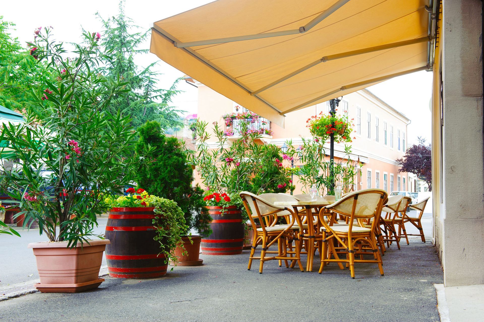 Outdoor café patio with wicker chairs, tables, potted plants, and a yellow canopy along a sunlit street