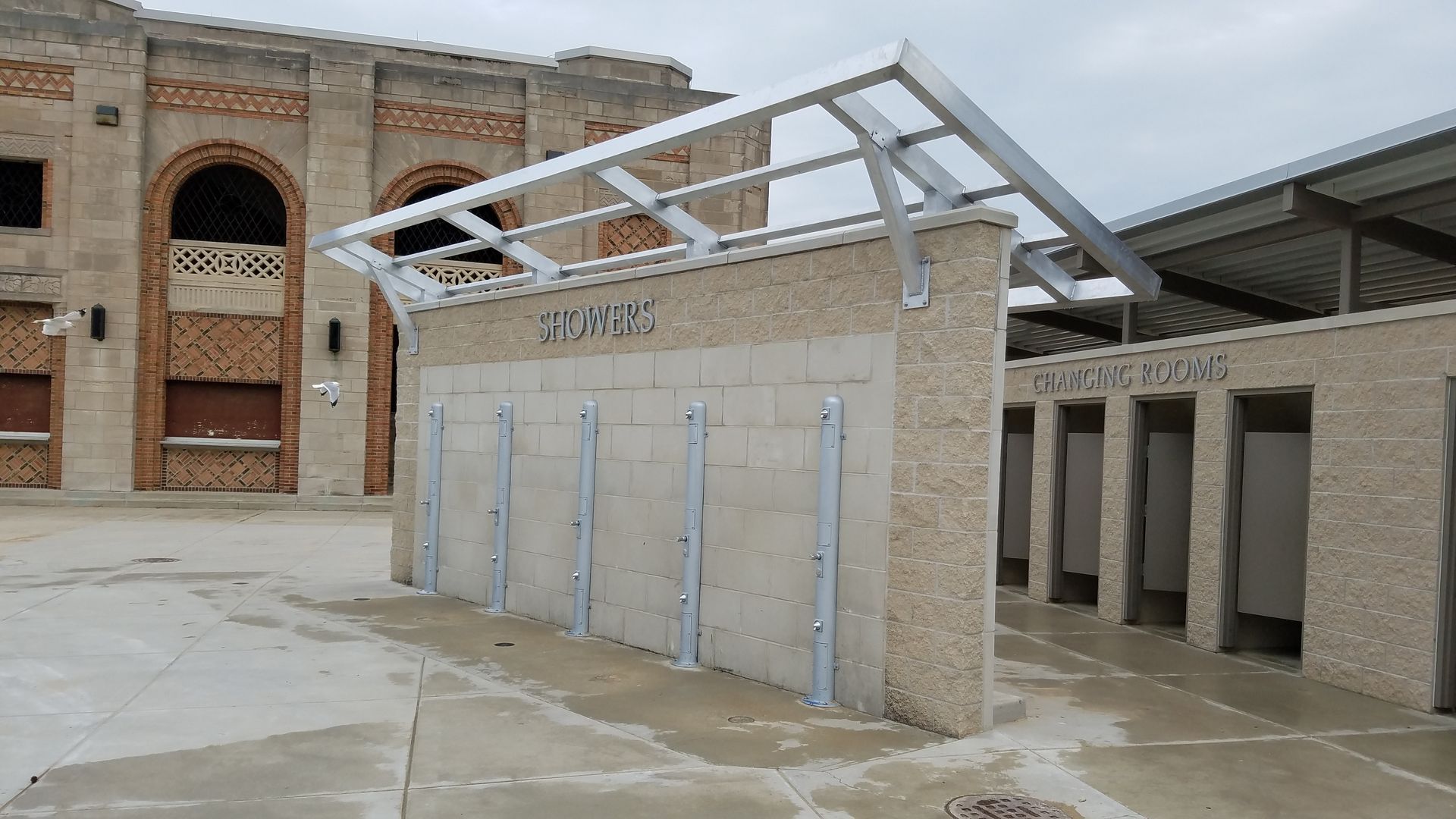 Outdoor concrete structure with metal roof frame and beige wall, in front of a brick building.