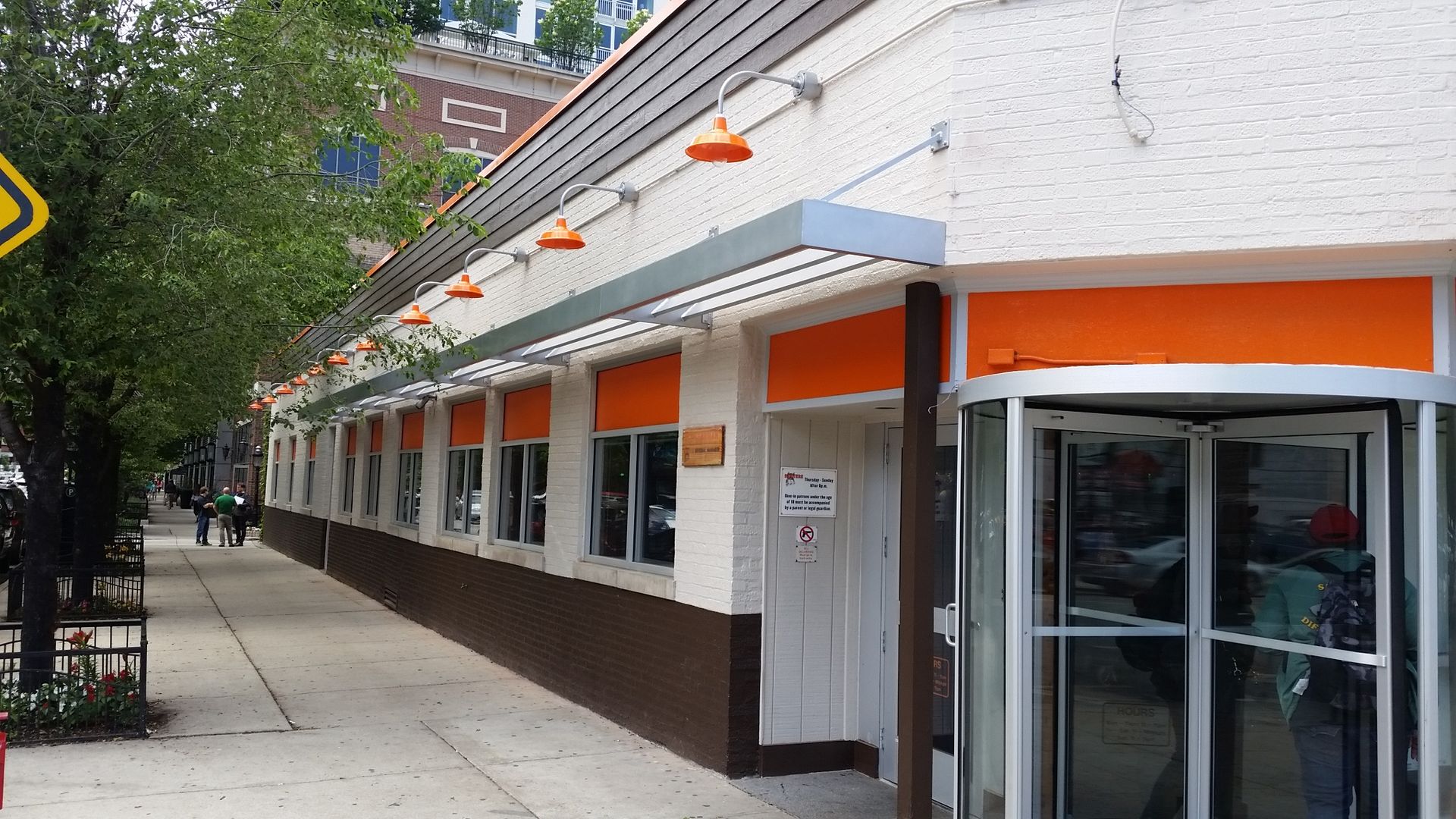 Long white storefront with orange awnings along a city sidewalk, viewed at an angle