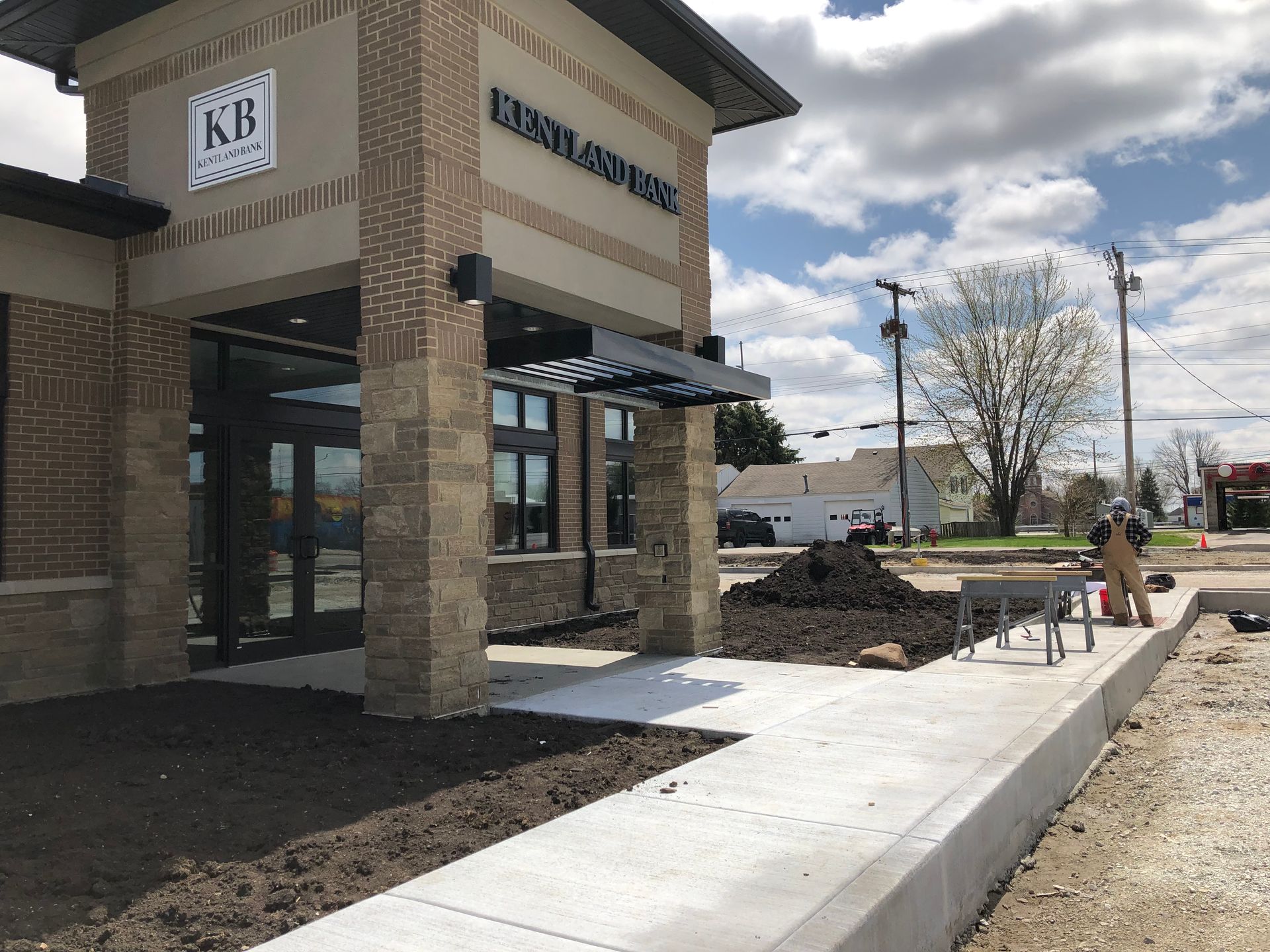Bank entrance with brick exterior, sidewalk, and a worker near a newly poured concrete curb under cloudy skies