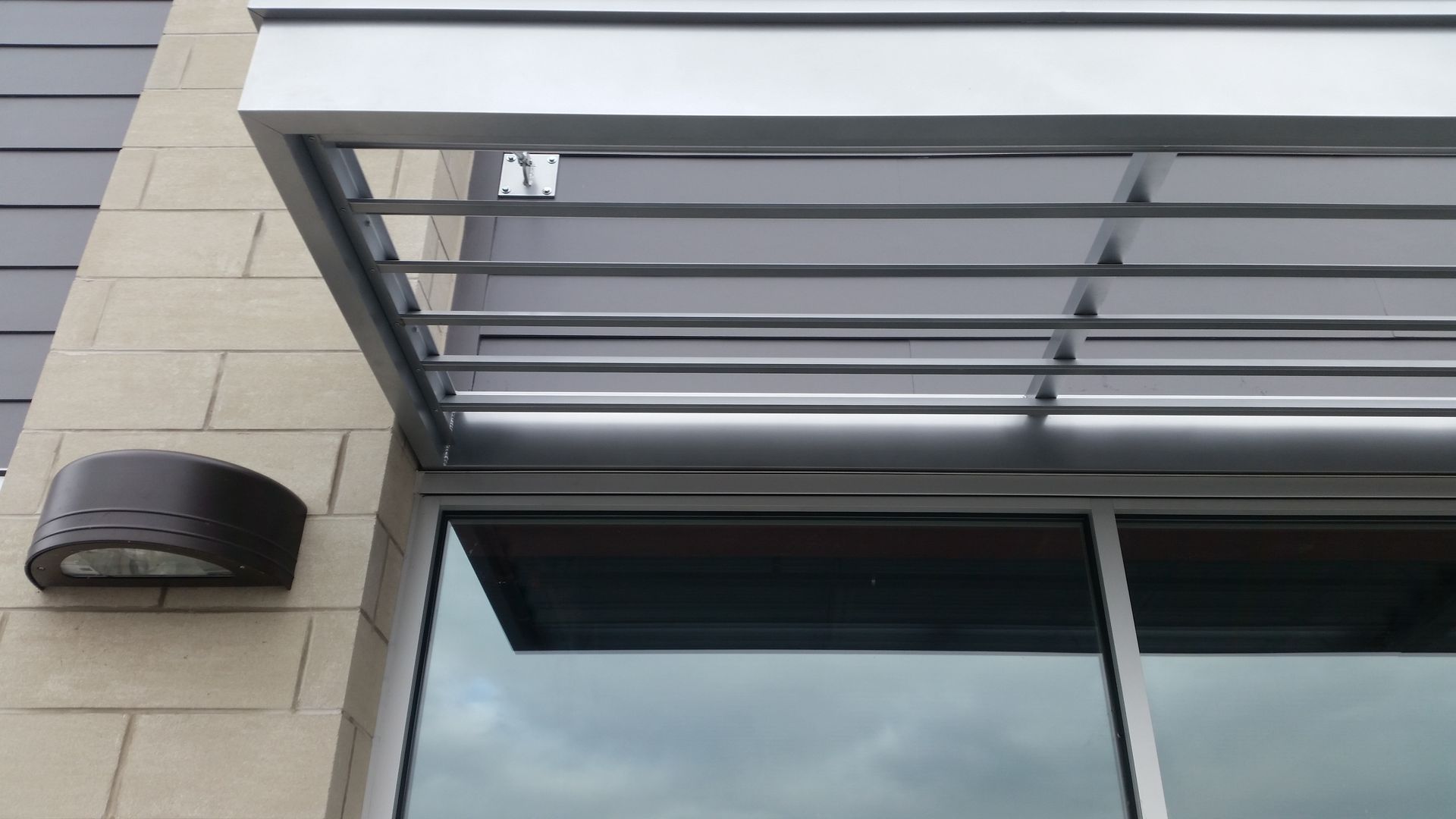 Modern building facade with a glass awning, horizontal beams, and a wall-mounted light.