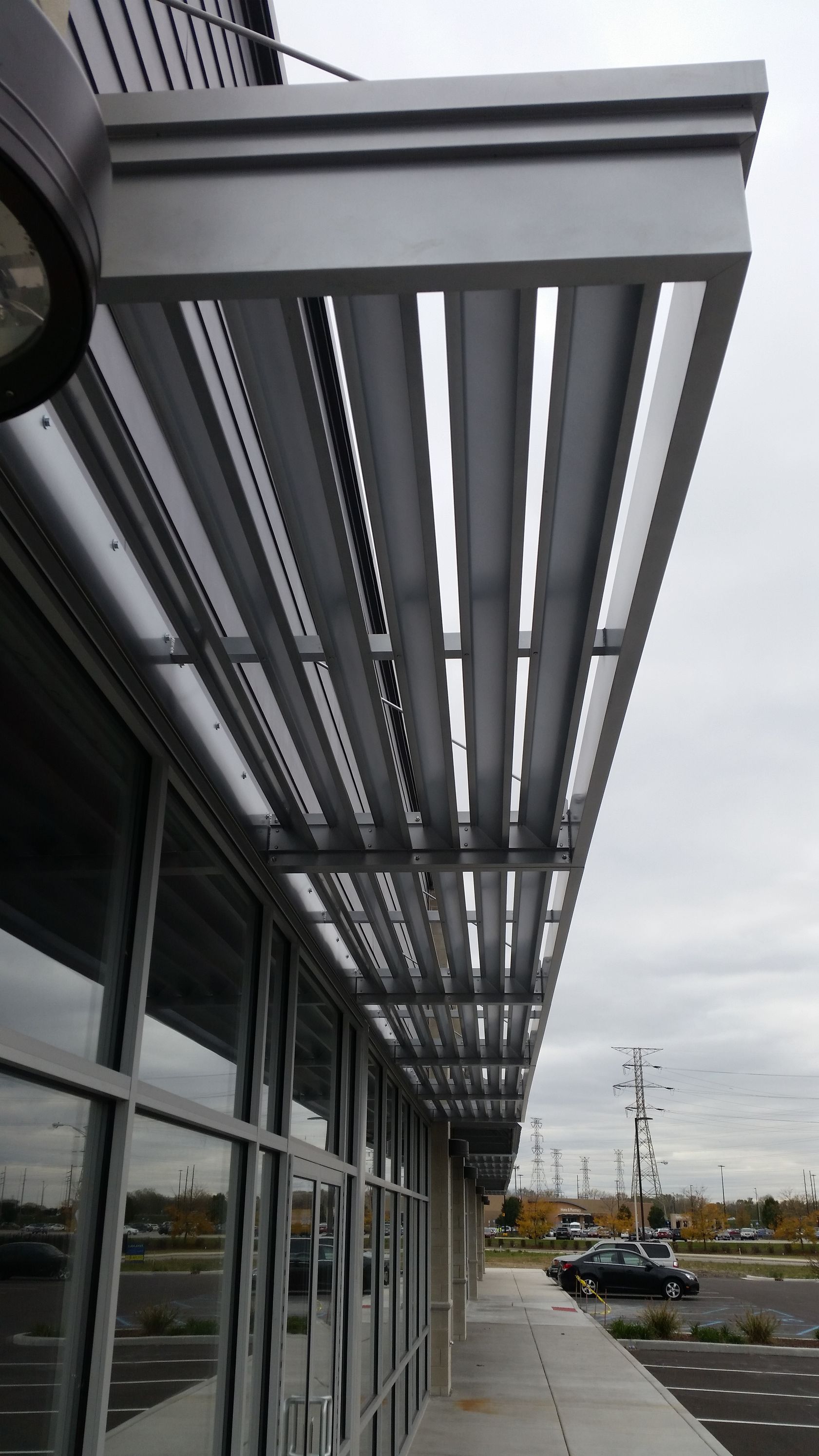 Modern storefront with glass windows and a metal awning extending over a parking lot.