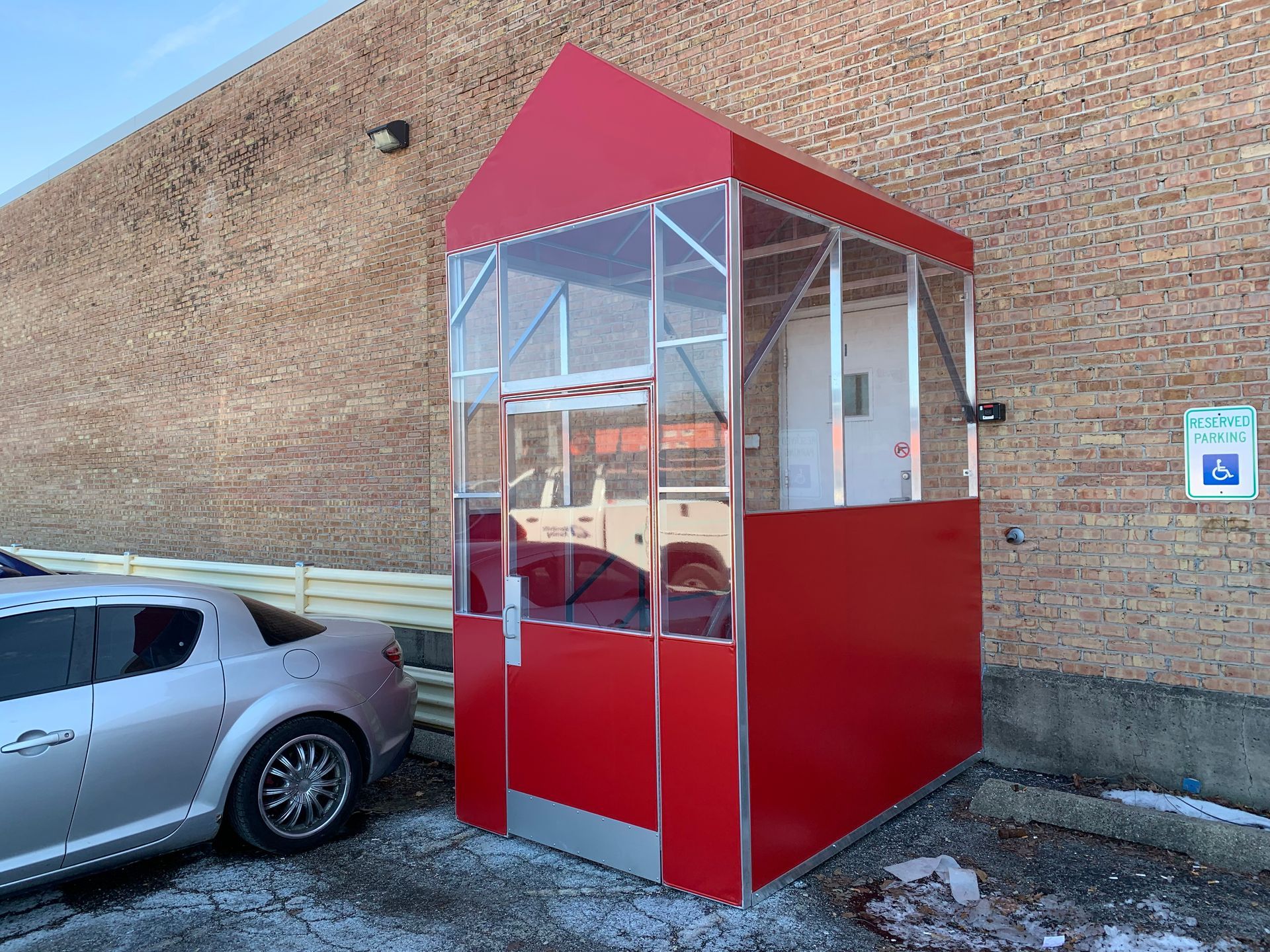 Red outdoor booth with clear panels beside a silver car against a brick wall