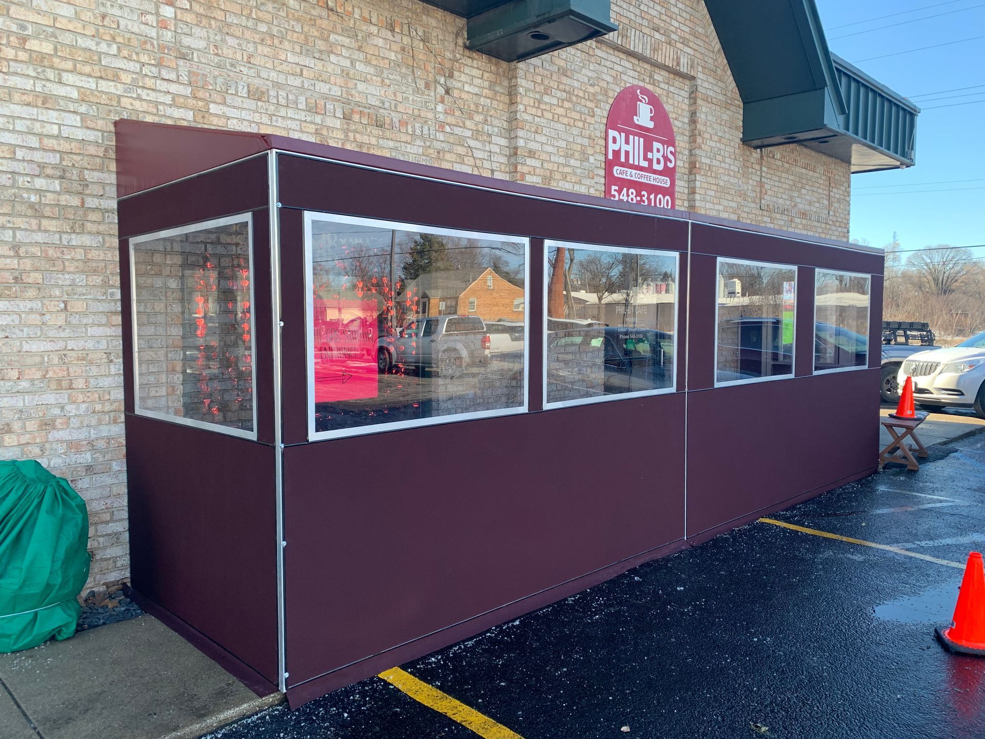 Maroon enclosed patio booth with windows outside a brick building, beside a parking lot and orange cone