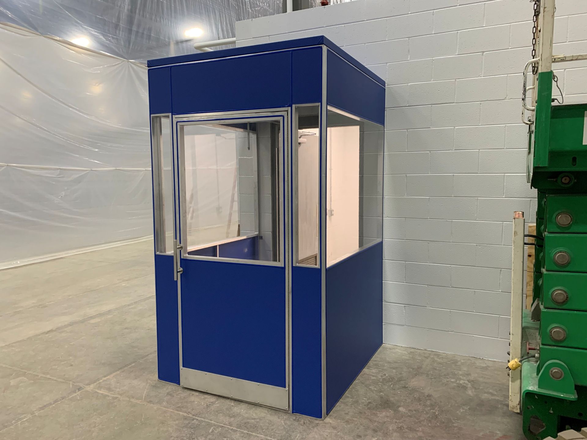 Blue industrial booth in a warehouse, with clear windows and a white brick wall behind it.